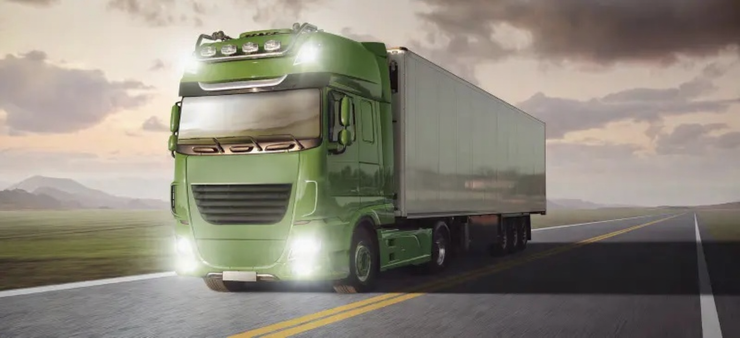 A green semi-truck driving down an open road with mountains in the background and cloudy skies above.