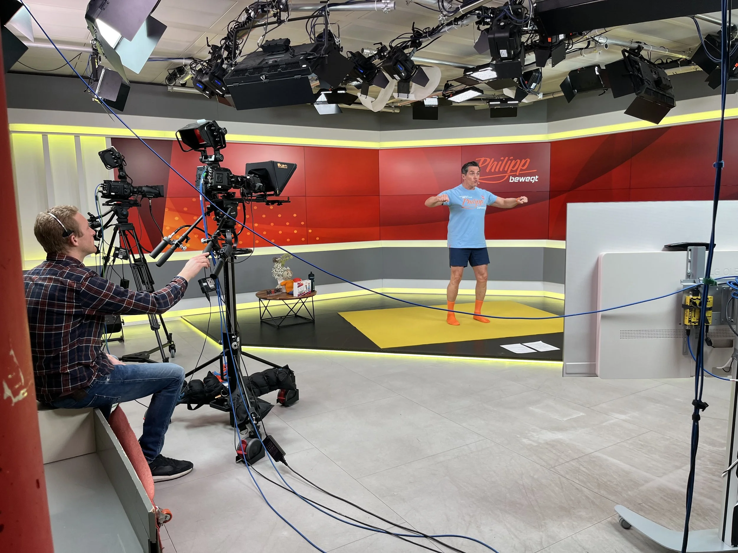 A man in a blue t-shirt and shorts standing on a yellow mat in a TV studio, with cameras and crew members filming him during a workout segment.