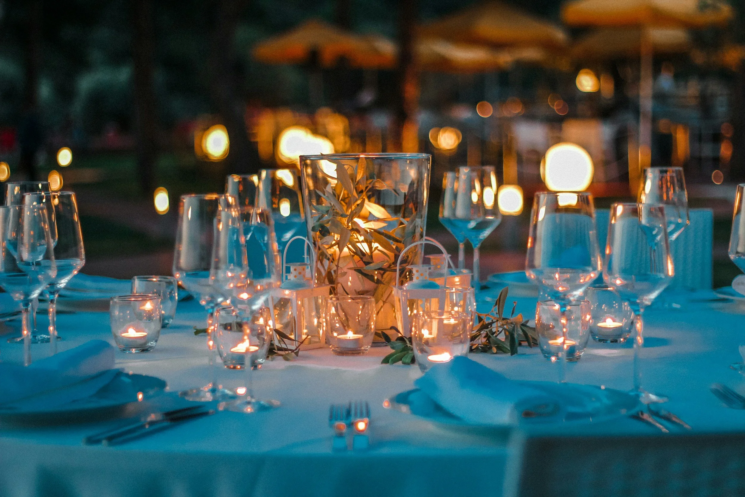 An elegant outdoor dinner table set with glassware, candles, and a centerpiece with foliage, illuminated by string lights at dusk.