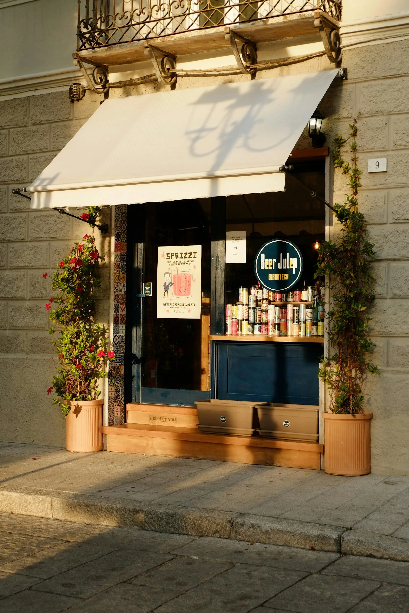 Facade of a small bar called 'Beer Julep Birroteca' with potted plants on each side, a large white awning, and a door with a poster. There are stacked cans on a shelf outside.