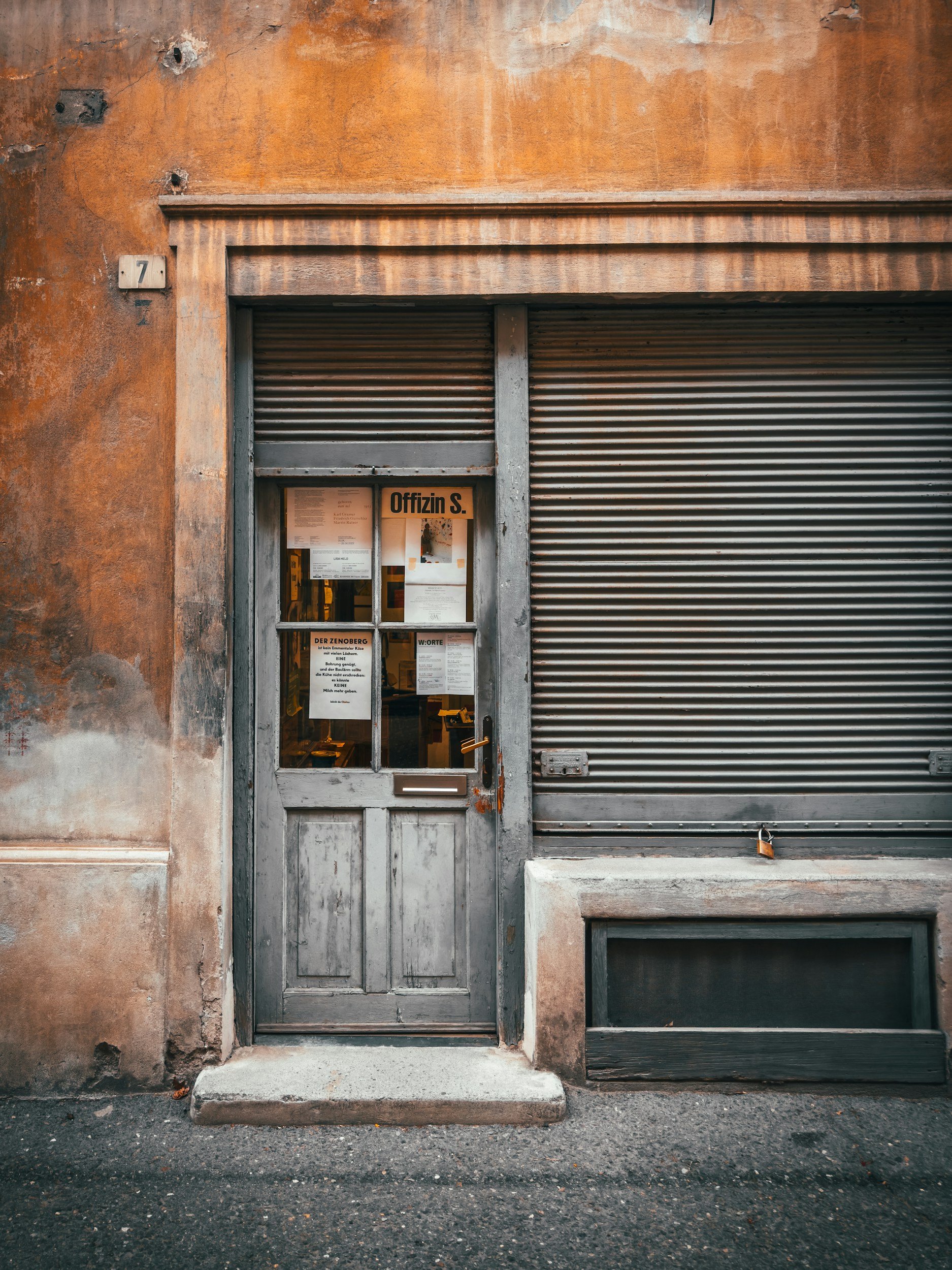 Old gray wooden door with notices and a sign 'Offizin S.' on glass window, set in a weathered brick wall with a closed metal rolling shutter beside it.