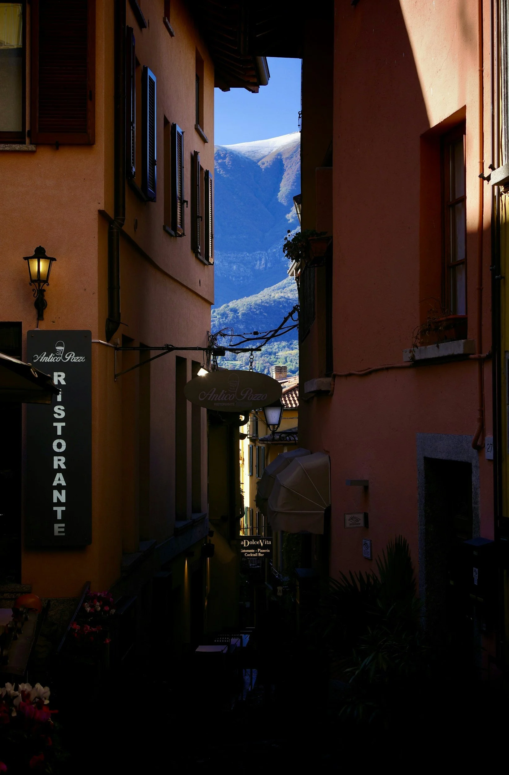 Narrow alleyway in an Italian town with colorful buildings, signs for a restaurant and bar, and mountains in the background.