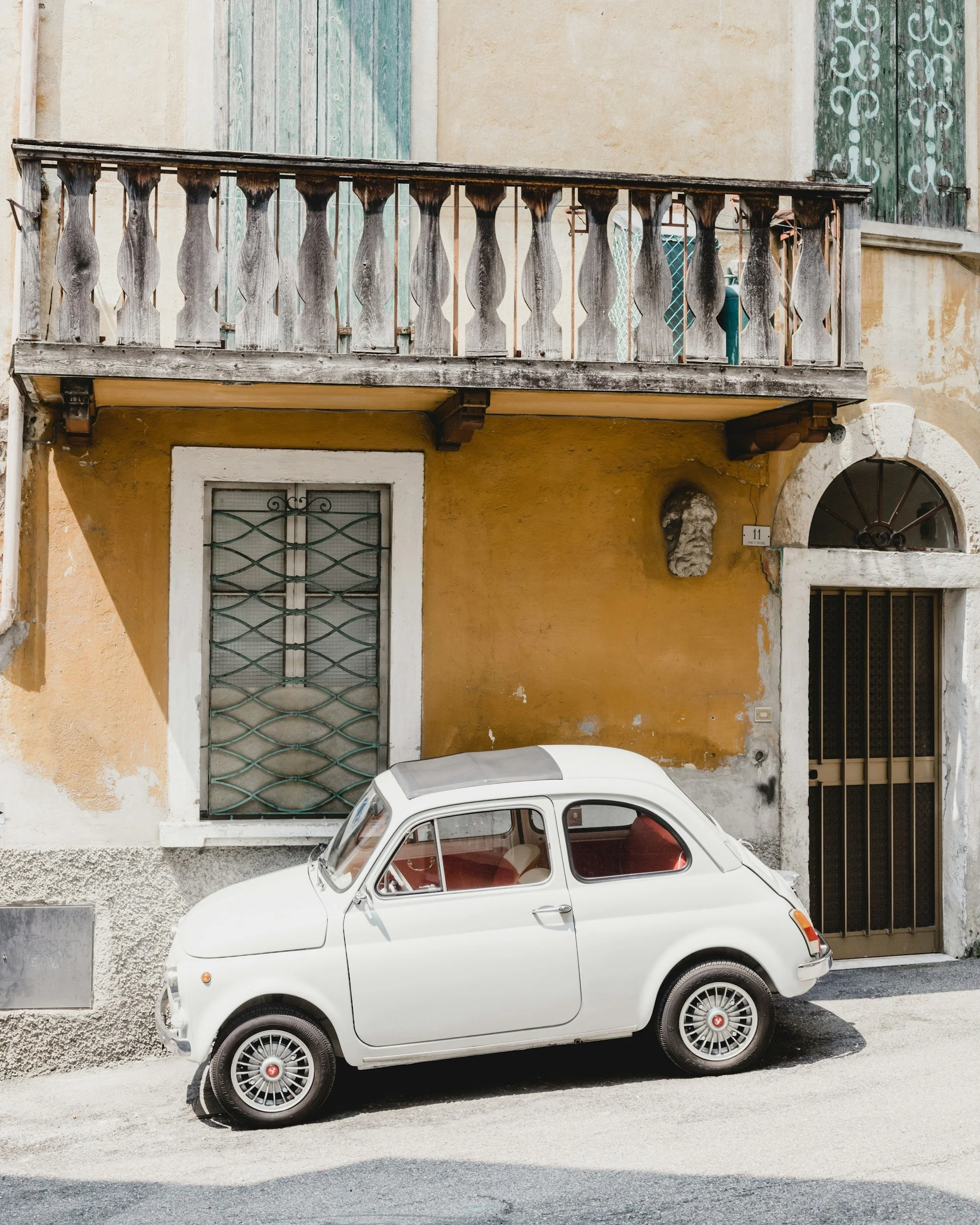 A white vintage Fiat 500 parked in front of a yellow and beige stucco building with a balcony and green-shuttered windows.