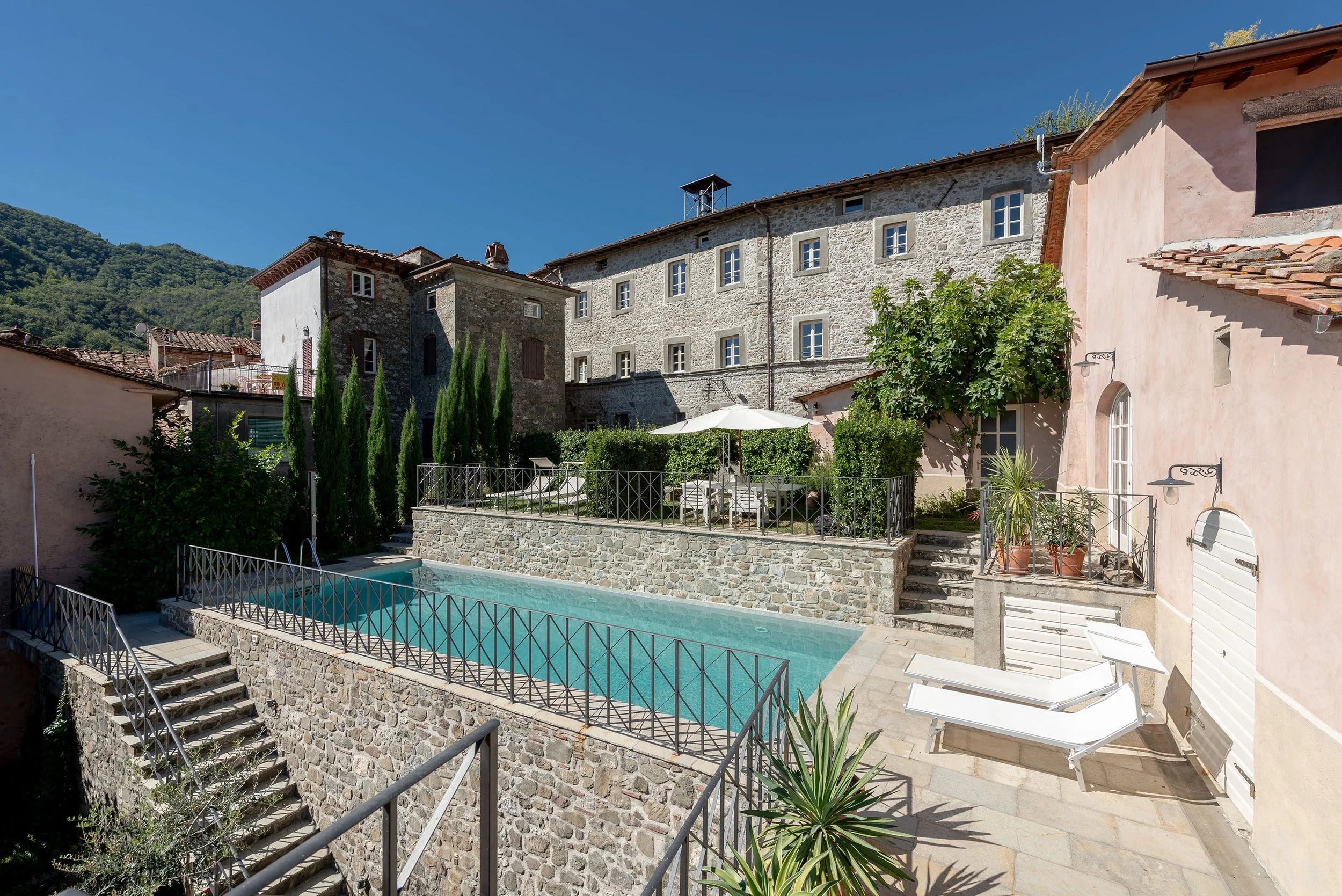 View of a stone and stucco hillside villa with a swimming pool, lounge chairs, umbrellas, potted plants, and outdoor seating area under a clear blue sky.