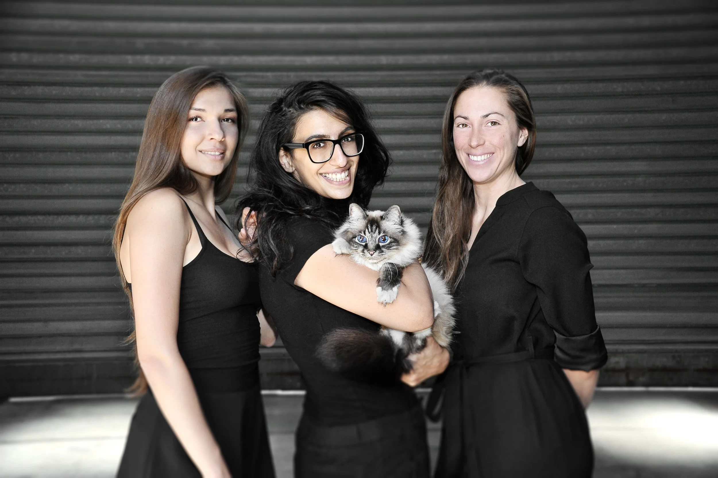 Three women and a kitten standing in front of a black roller door, with the woman in the middle holding the kitten, all smiling at the camera.