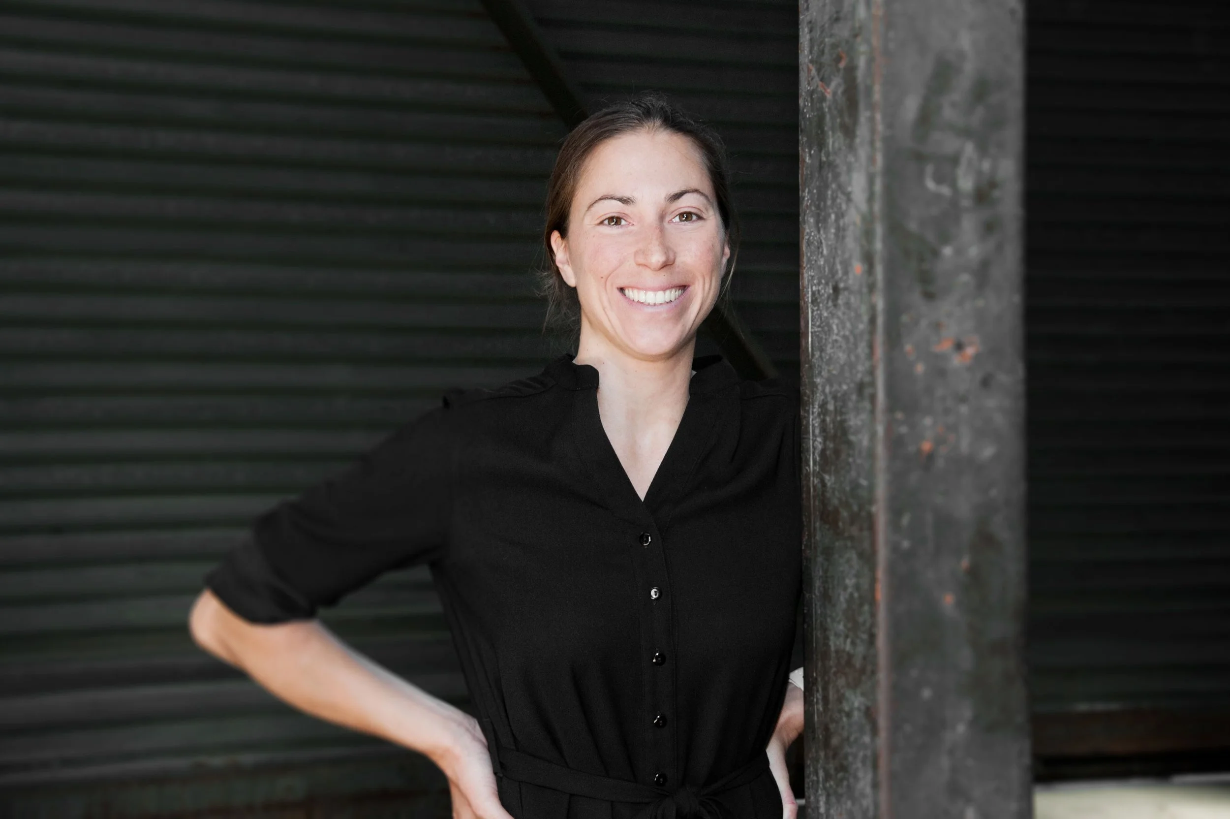 A woman with brown hair wearing a black shirt, smiling and standing with hands on her hips, partly behind a rusty metal post in front of a dark, ribbed background.