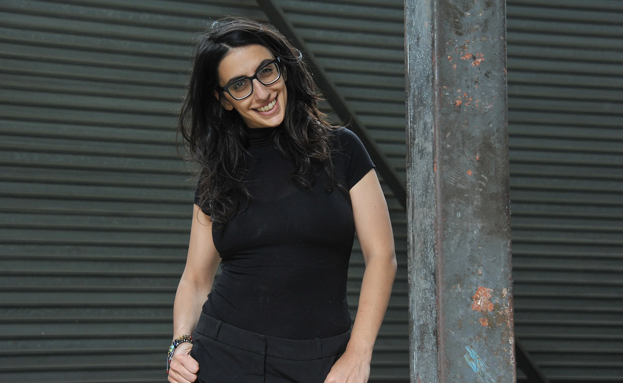 A woman with long dark hair and glasses wearing a black turtleneck shirt and black pants, smiling and standing in front of a metallic background and a rusted metal post.