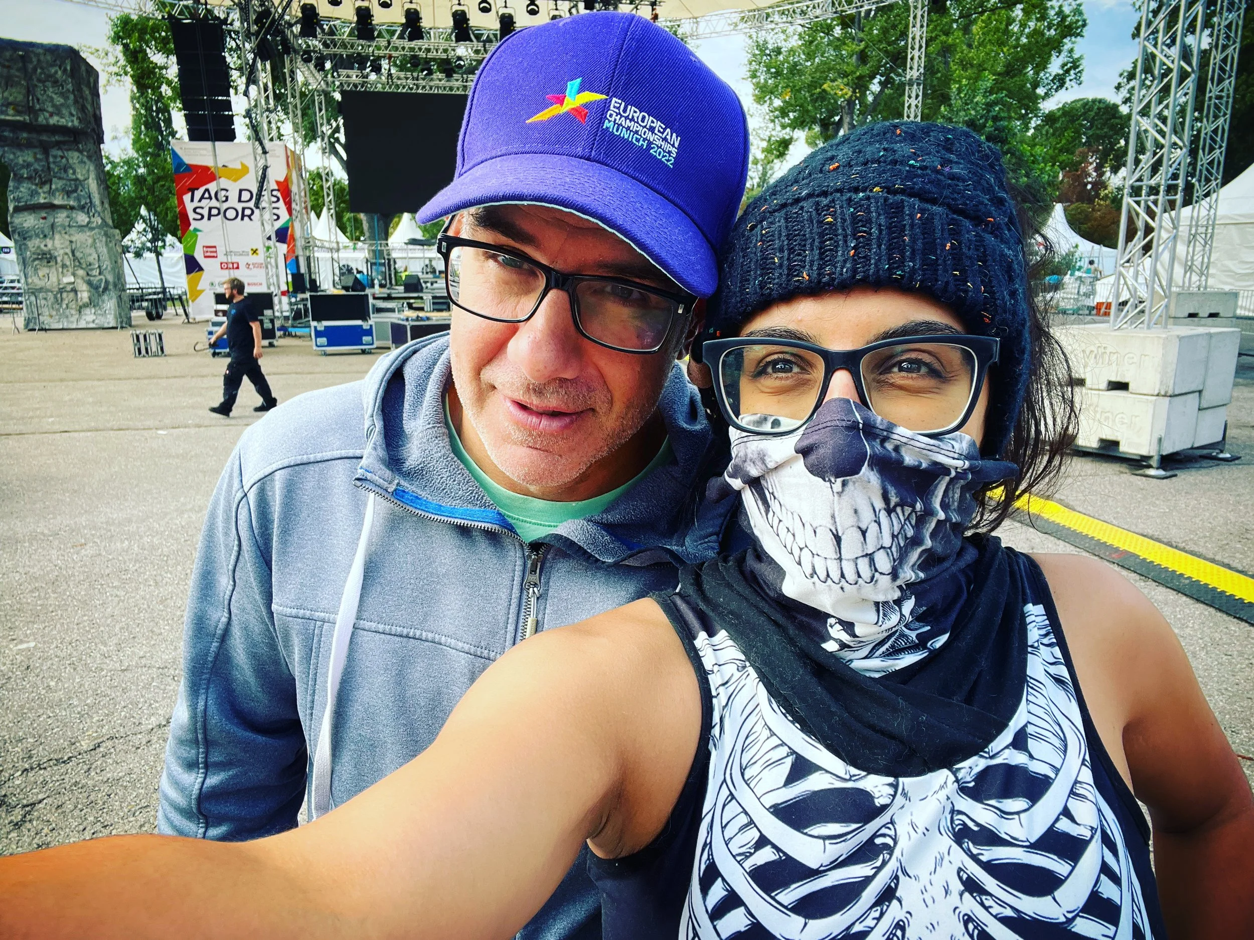 A man and a woman taking a selfie at an outdoor event. The man is wearing a blue cap and glasses, while the woman has a beanie, glasses, and a skeleton-themed face mask and top. In the background, there is a stage, trees, and event equipment.