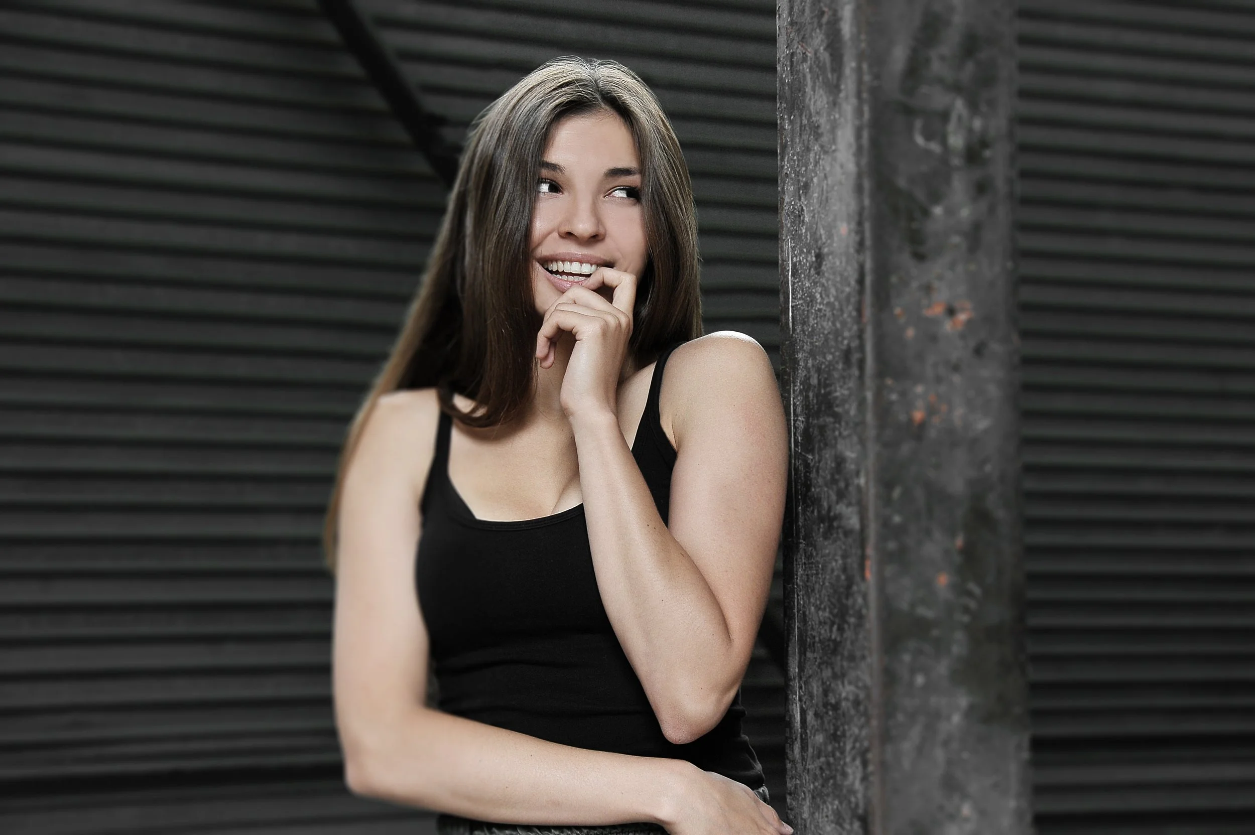 A young woman with long brown hair in a black tank top, smiling playfully, with her finger on her lips, standing against a dark, industrial background.