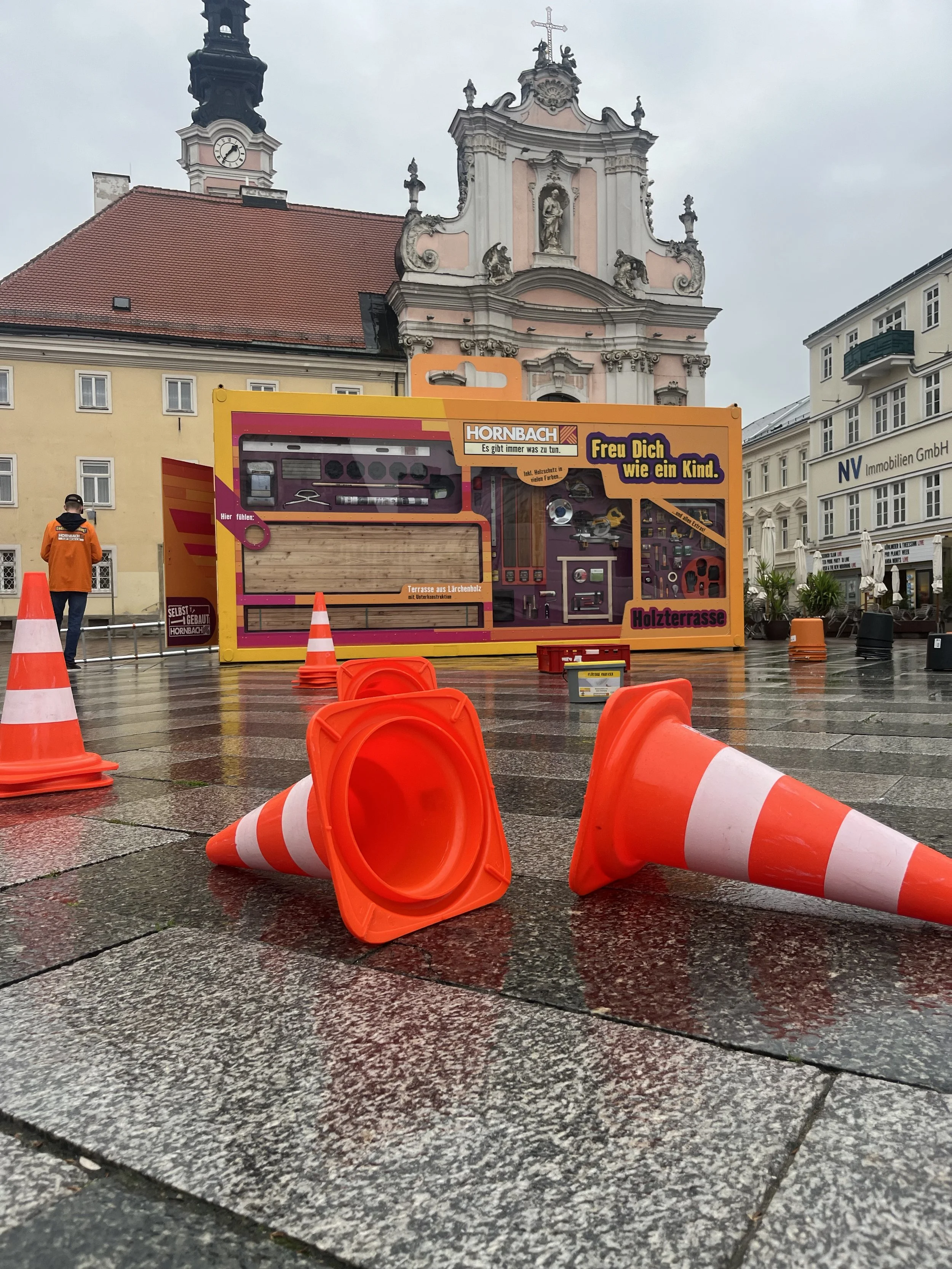 Construction site on wet pavement with orange traffic cones, a man in orange jacket, and a colorful decorated large box structure resembling a toy or promotional display in front of historic buildings and a cloudy sky.