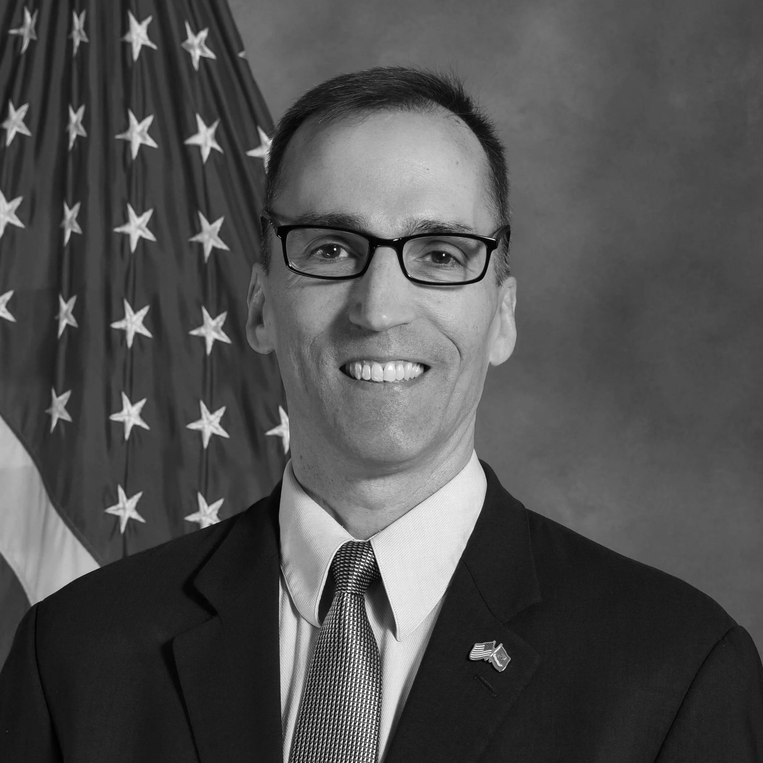 Smiling man wearing glasses and a dark suit in front of a United States flag.