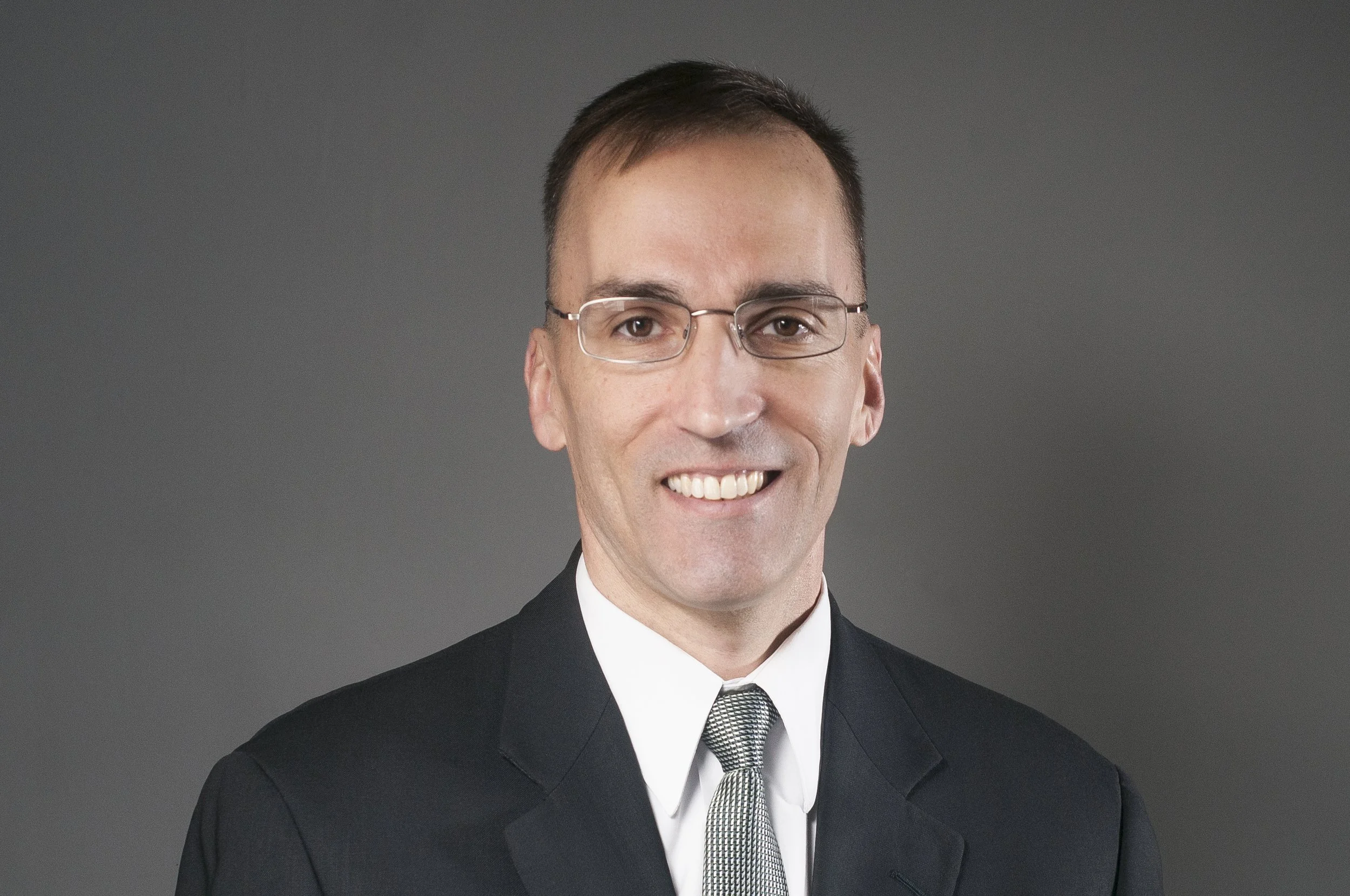 Dr. Gary J. Sampson headshot. Man wearing glasses in a dark suit with a green patterned tie.