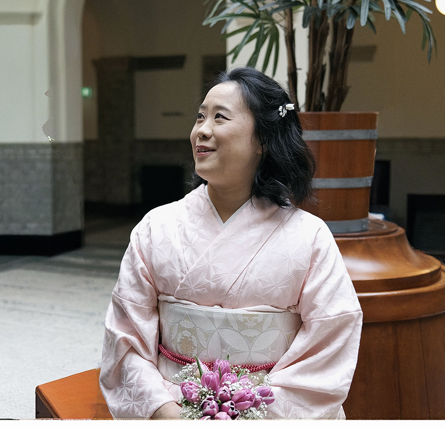 A woman in a light pink kimono holding a pink flower bouquet sits indoors near a large plant.