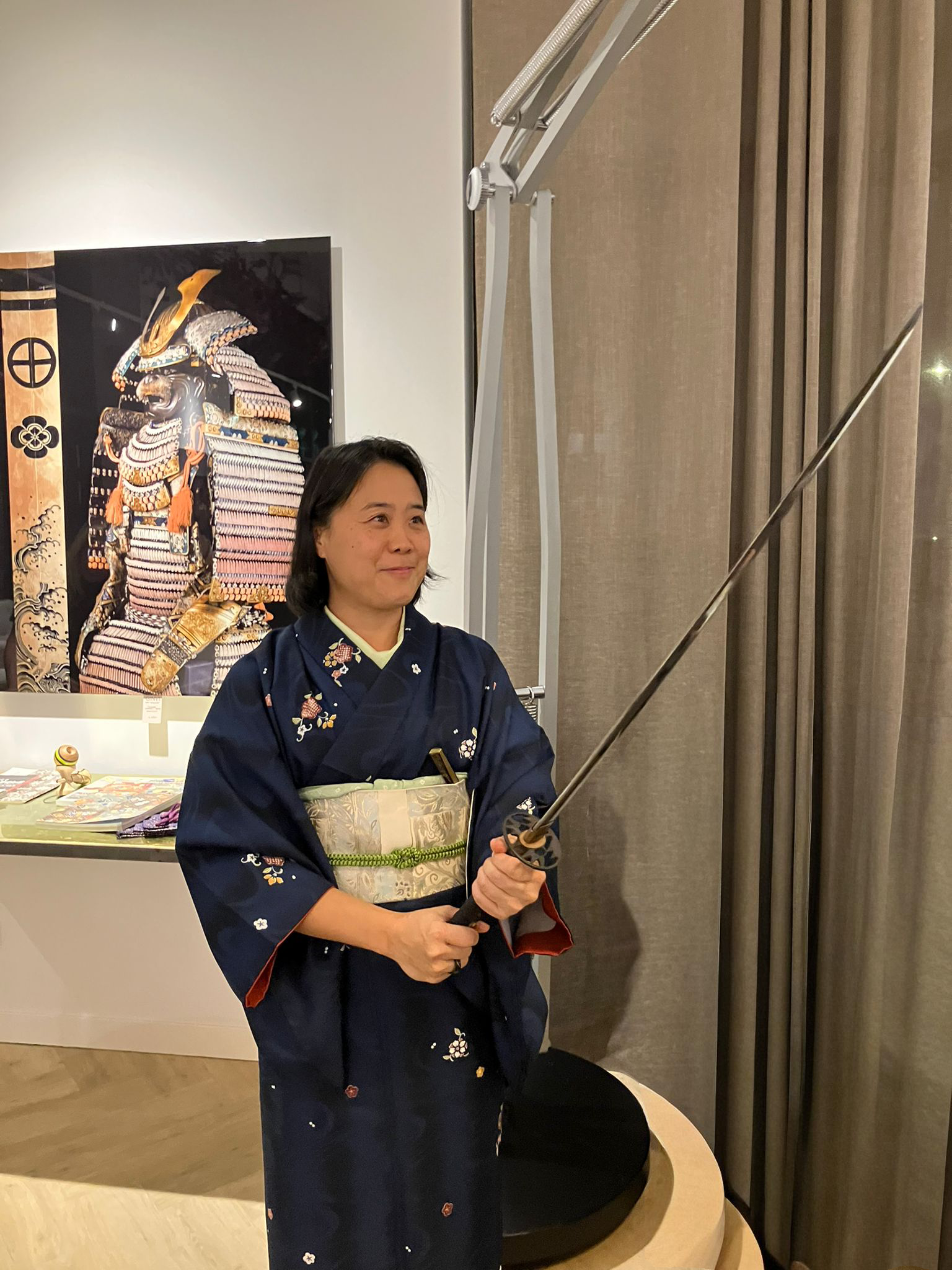 A woman wearing a traditional dark blue Japanese kimono is holding a katana sword with a black handle. She is standing indoors near beige curtains and a display with artwork or artifacts in the background.