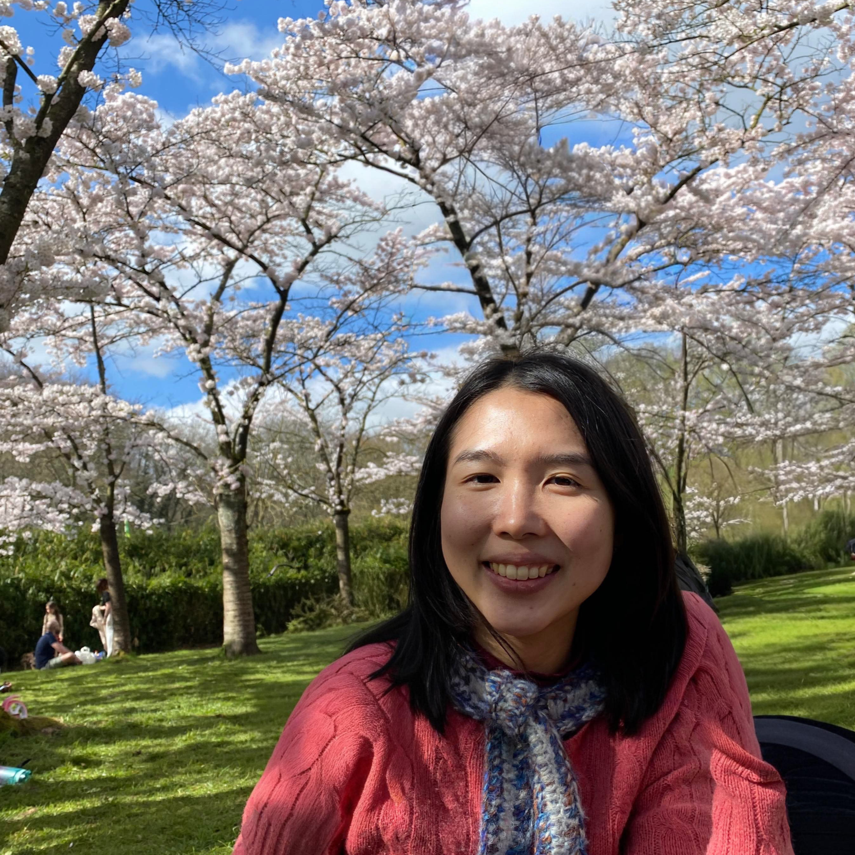 A woman with black hair smiling at the camera in a park with blooming cherry blossom trees and a blue sky with clouds.