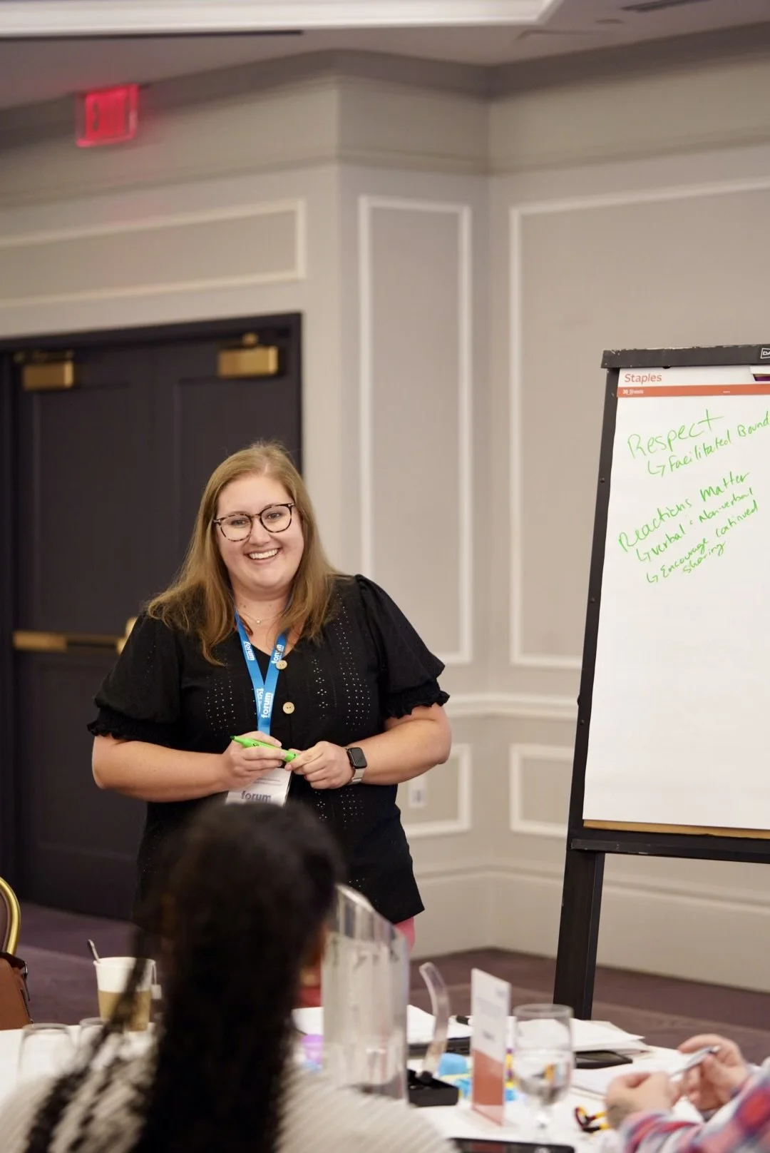 A woman with glasses, wearing a black top and a conference badge, is smiling while standing in front of a whiteboard, holding a green marker in a conference room.