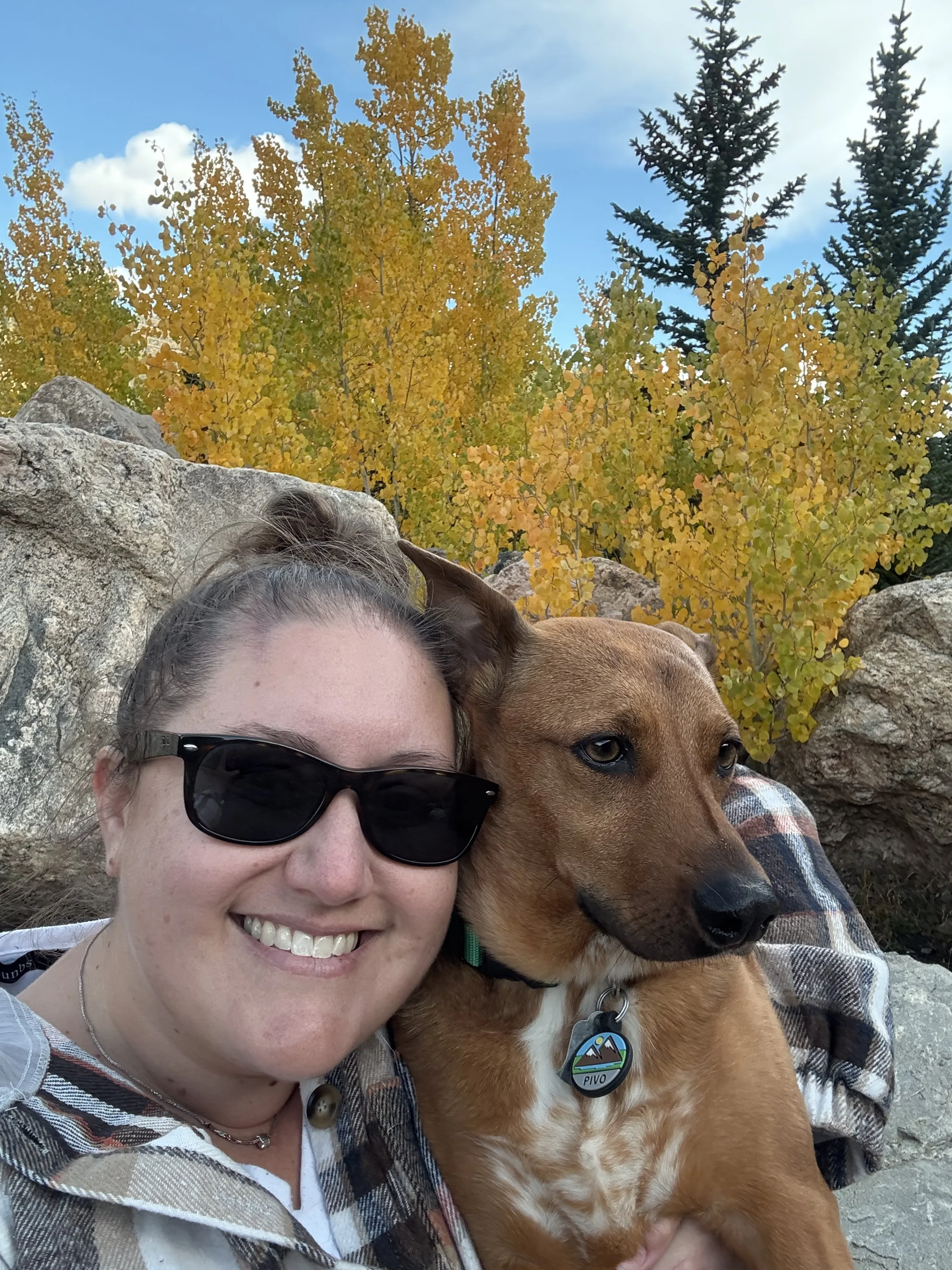 A smiling woman wearing sunglasses and a plaid shirt, holding a brown dog with a collar and tag, with yellow and green trees and rocks in the background.