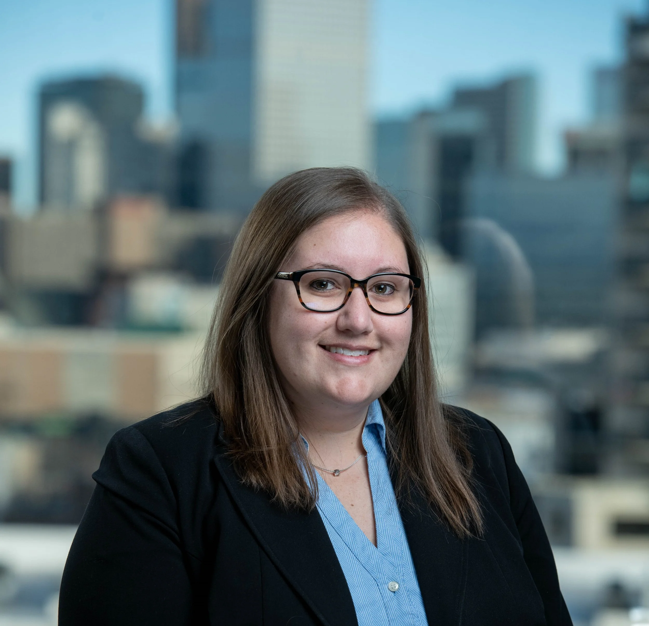Professional woman with glasses and brown hair, wearing a black blazer and blue blouse, smiling against a city skyline background.