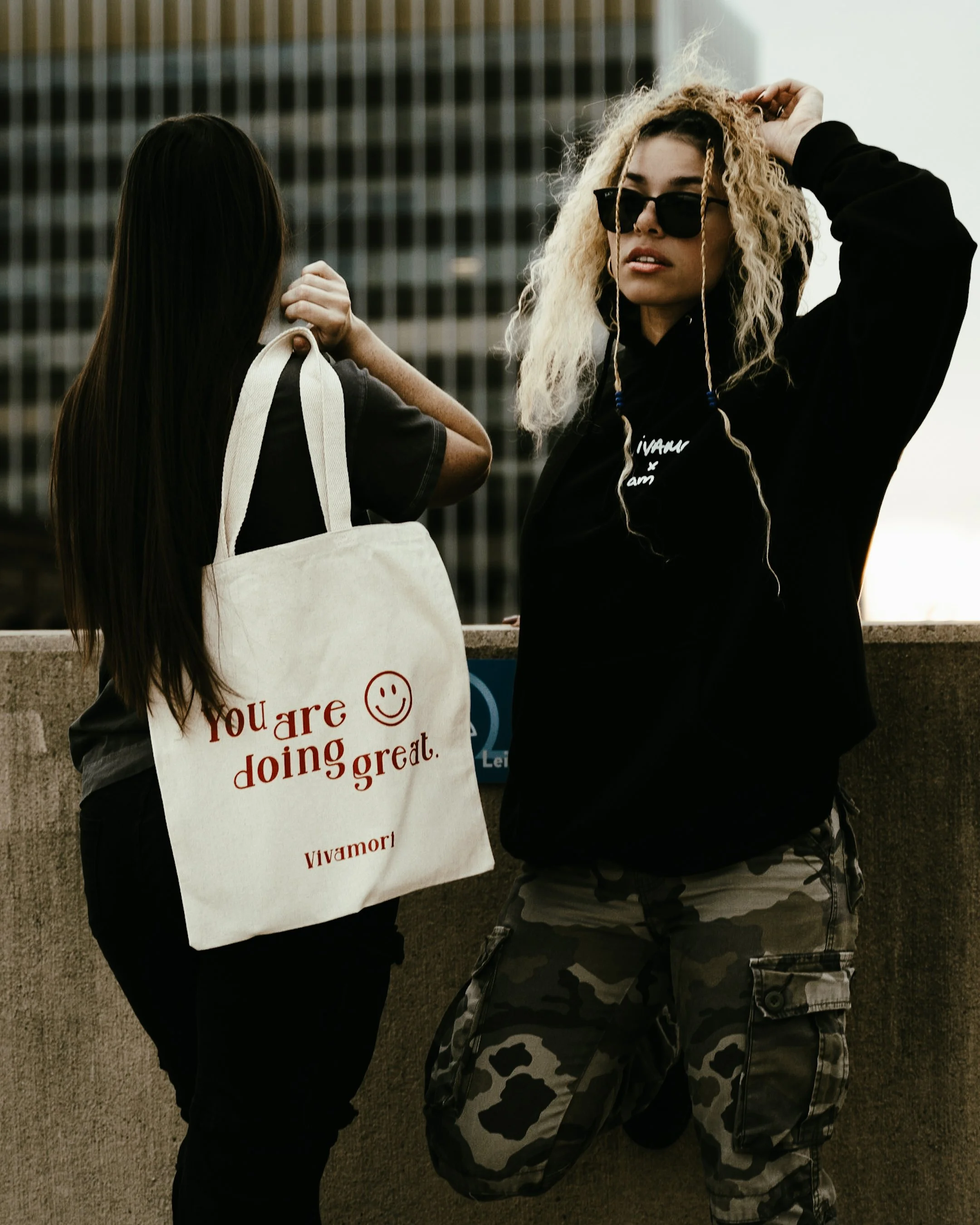 Two women talking outdoors, one holding a tote bag that says "You are doing great" with a smiley face and the brand name Vivamori, in front of a cityscape background.