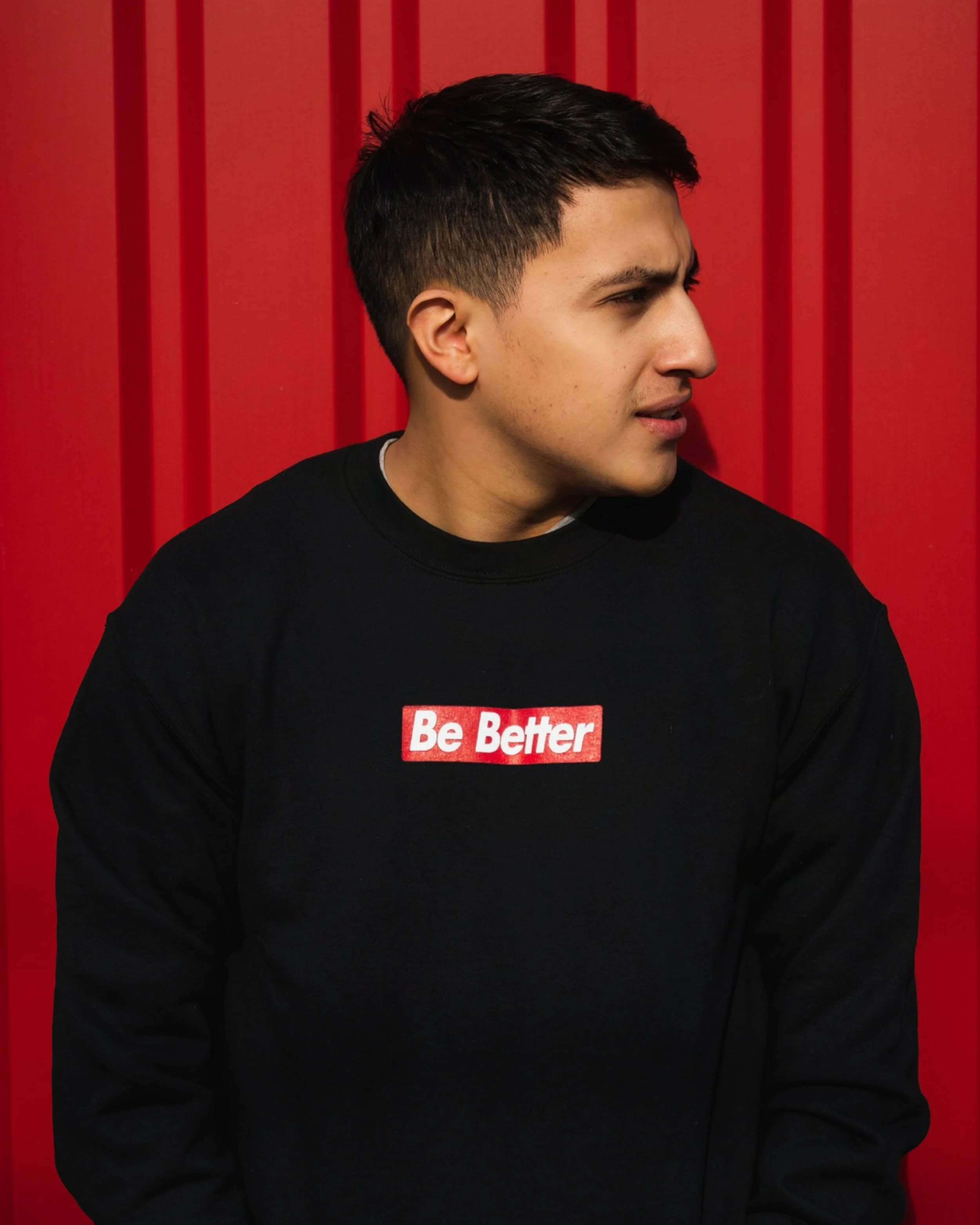 A young man with dark hair, wearing a black sweatshirt with the words 'Be Better' on it, looking to the right against a red background.