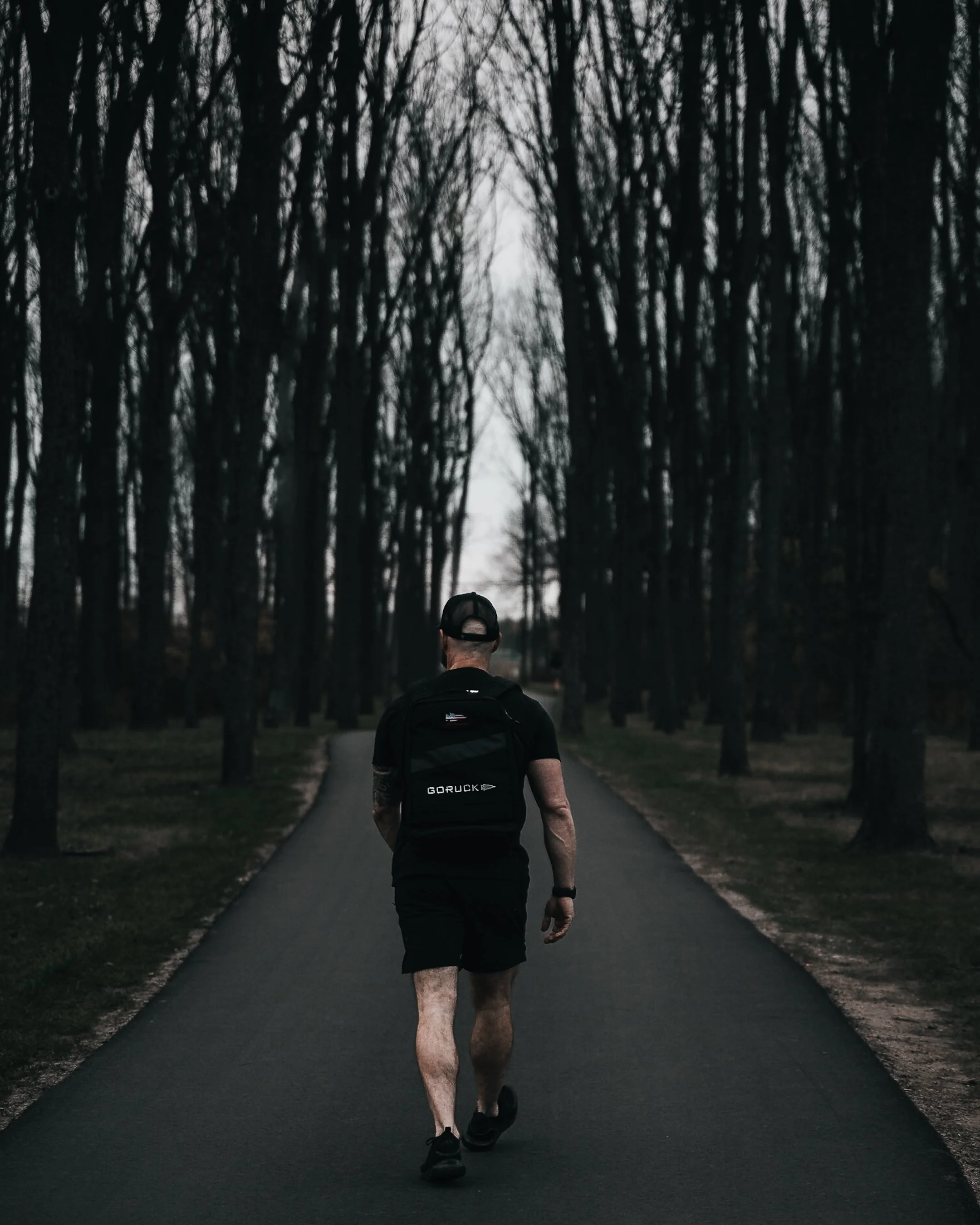 A man walking alone on a paved path through a park with leafless trees on both sides, during late evening or early night.