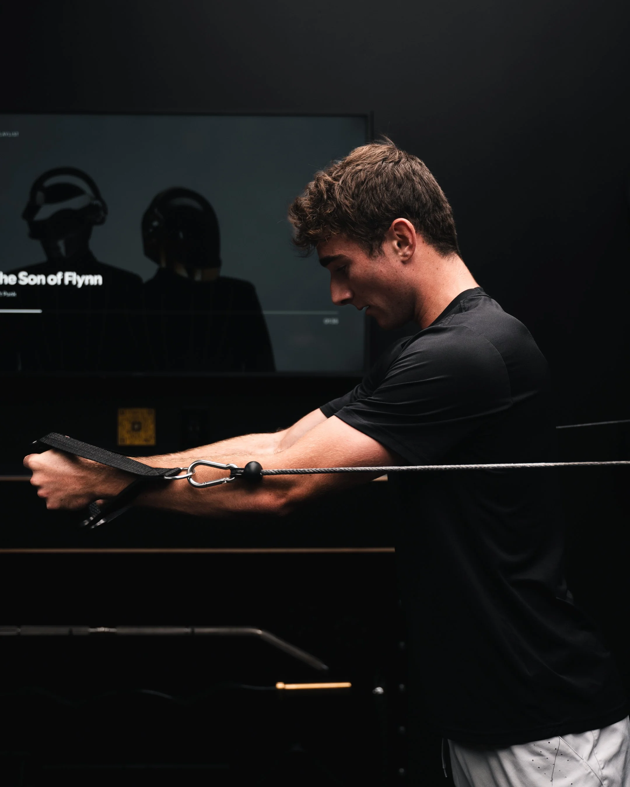 Young man exercising with a resistance band in a gym, black background, TV screen showing headphones and music title.