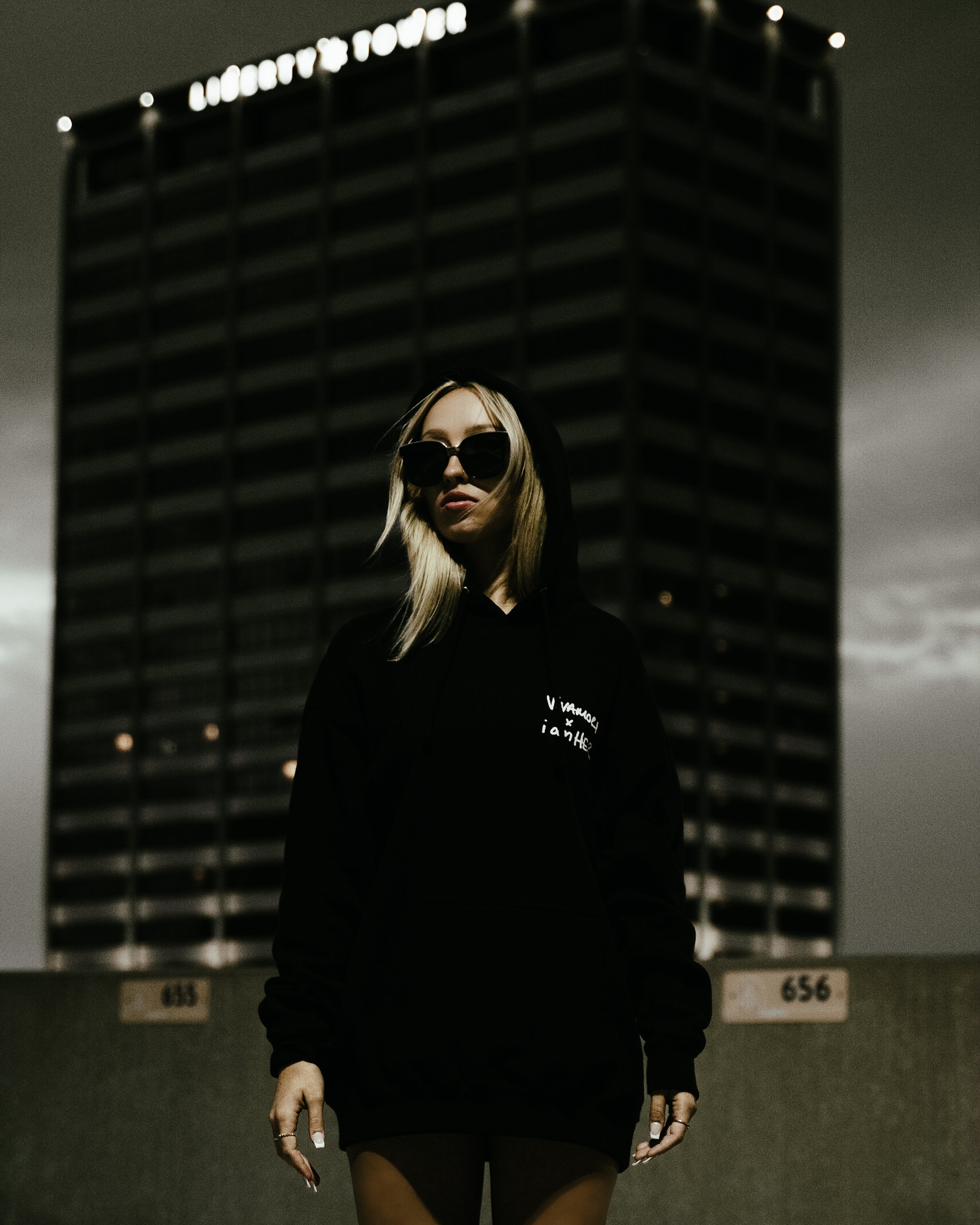 A woman wearing sunglasses and a black hoodie standing in front of a tall building at night.