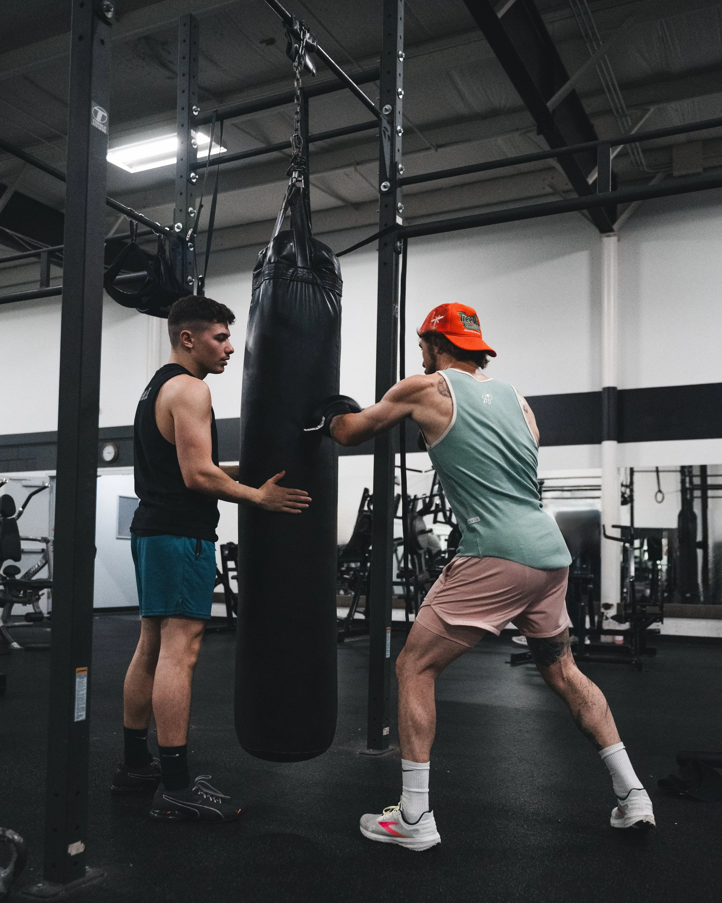 Two men hit a punching bag in a gym. One man is giving instructions, the other is wearing a tank top, shorts, a red hat, and boxing gloves.