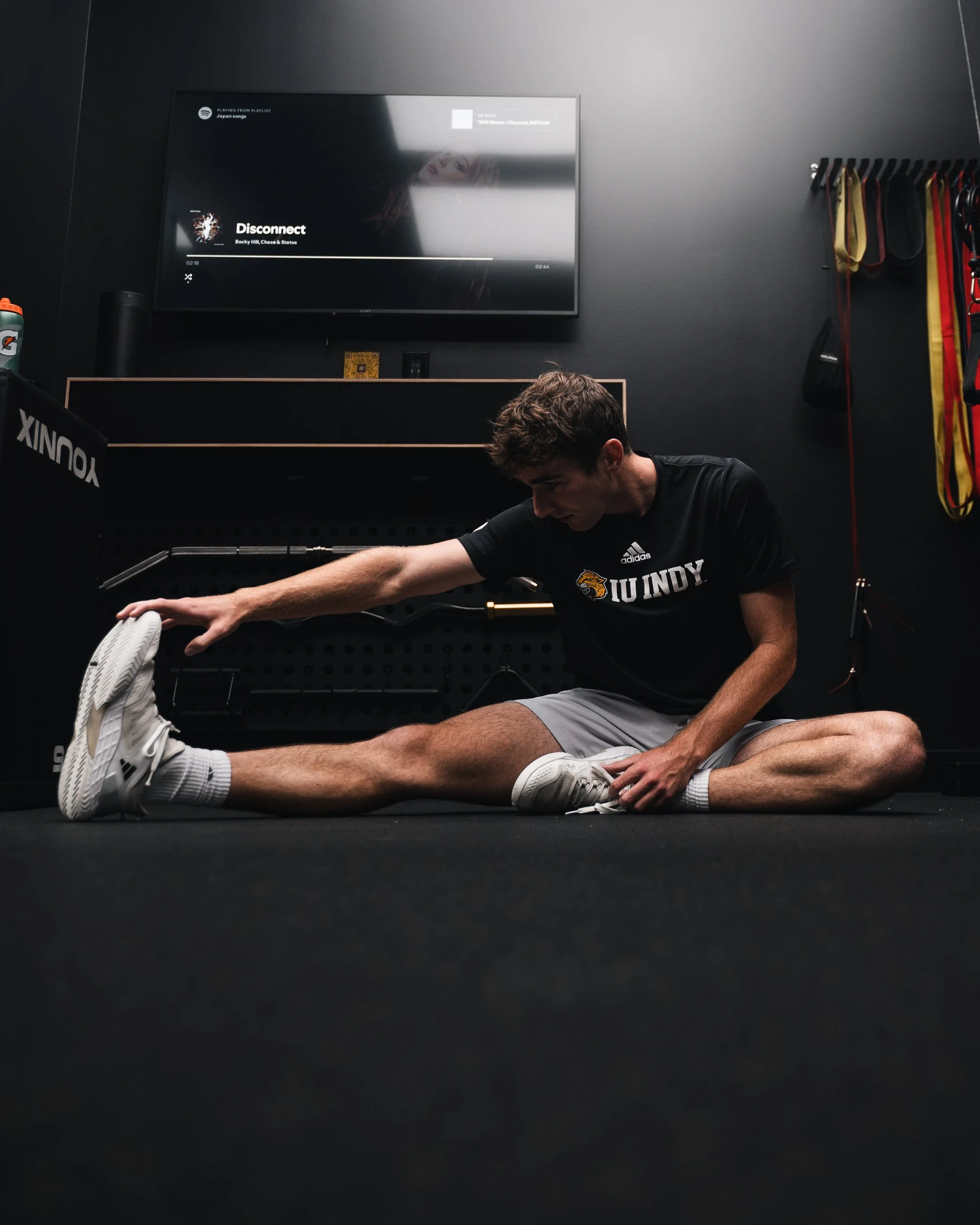 A man stretching on the floor of a gym, wearing a black Indiana University shirt and gray shorts, with a television mounted on the wall behind him displaying a Spotify music player.
