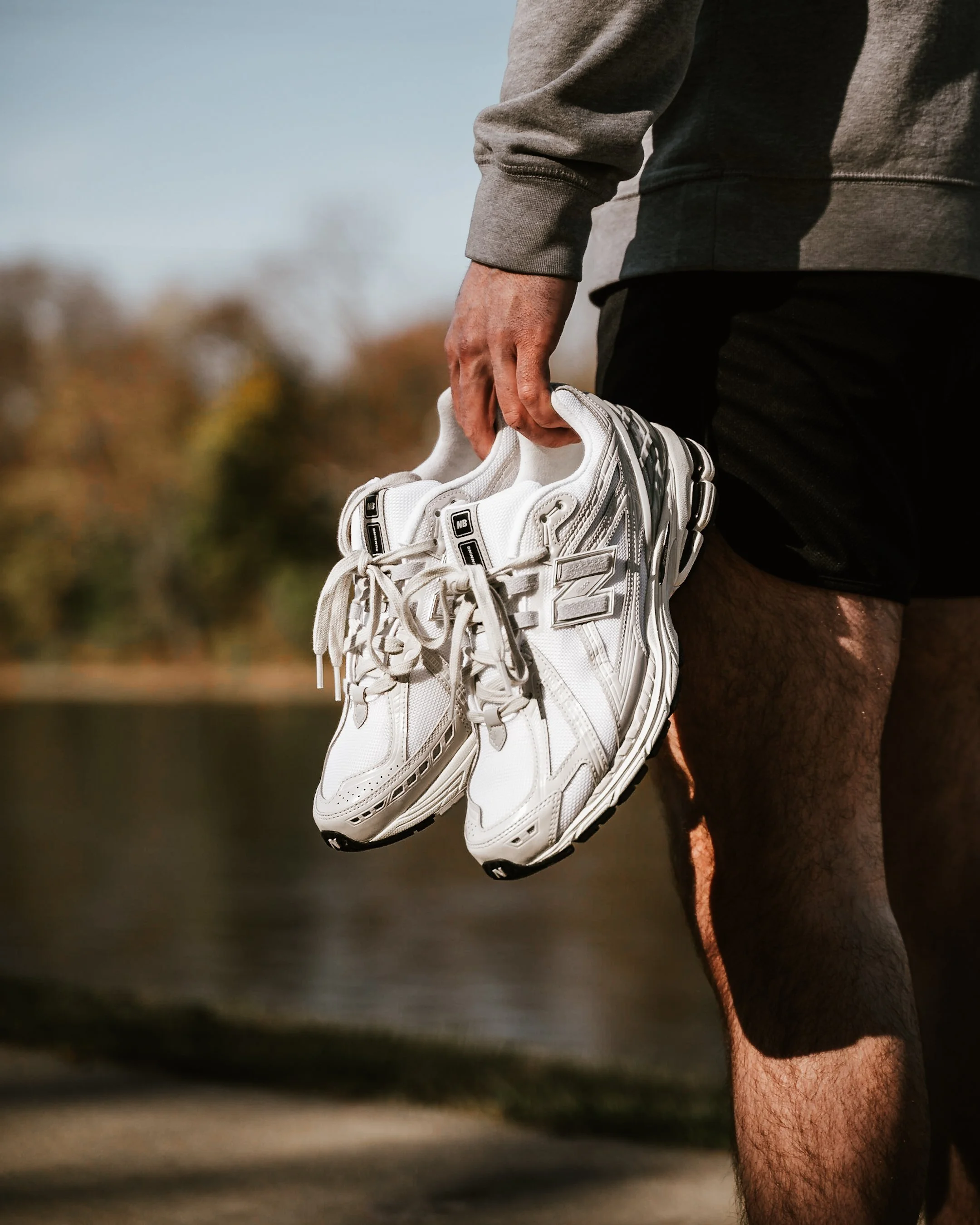 A person holding a pair of white athletic shoes outdoors, near a body of water with blurred autumn trees in the background.