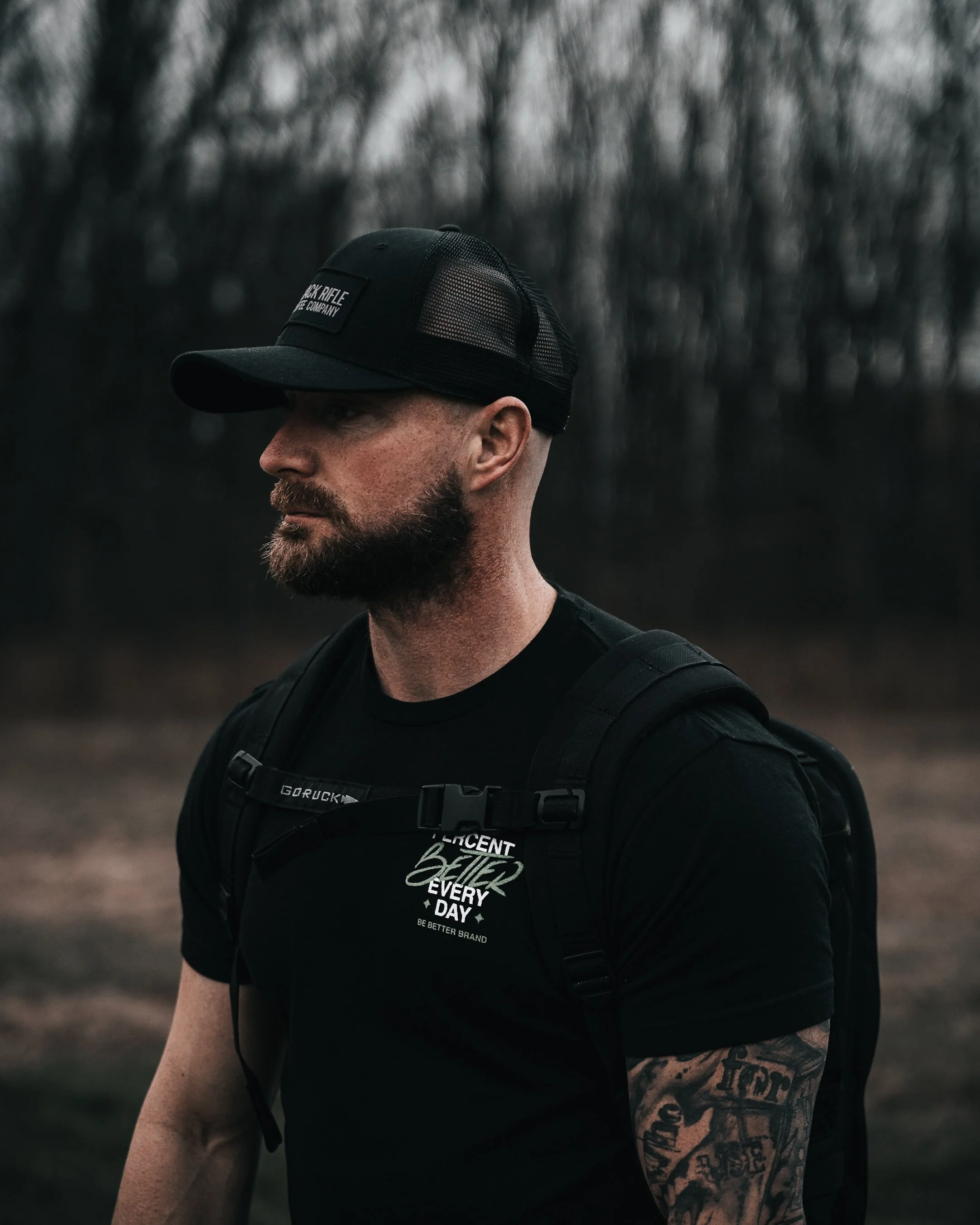 A man with a beard wearing a black cap and black t-shirt with white text, carrying a black backpack, outdoors near a forest with leafless trees.