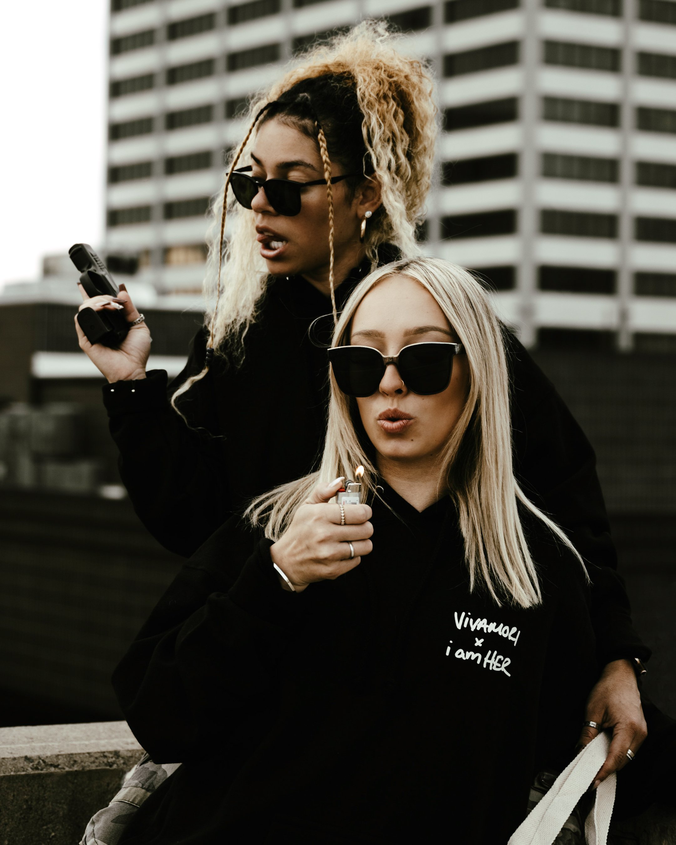 Two women with black sunglasses standing outdoors in front of a modern building, one with blonde hair and the other with curly, blonde and brown hair, both wearing dark clothing, holding lighters and a small object, possibly a cell phone.