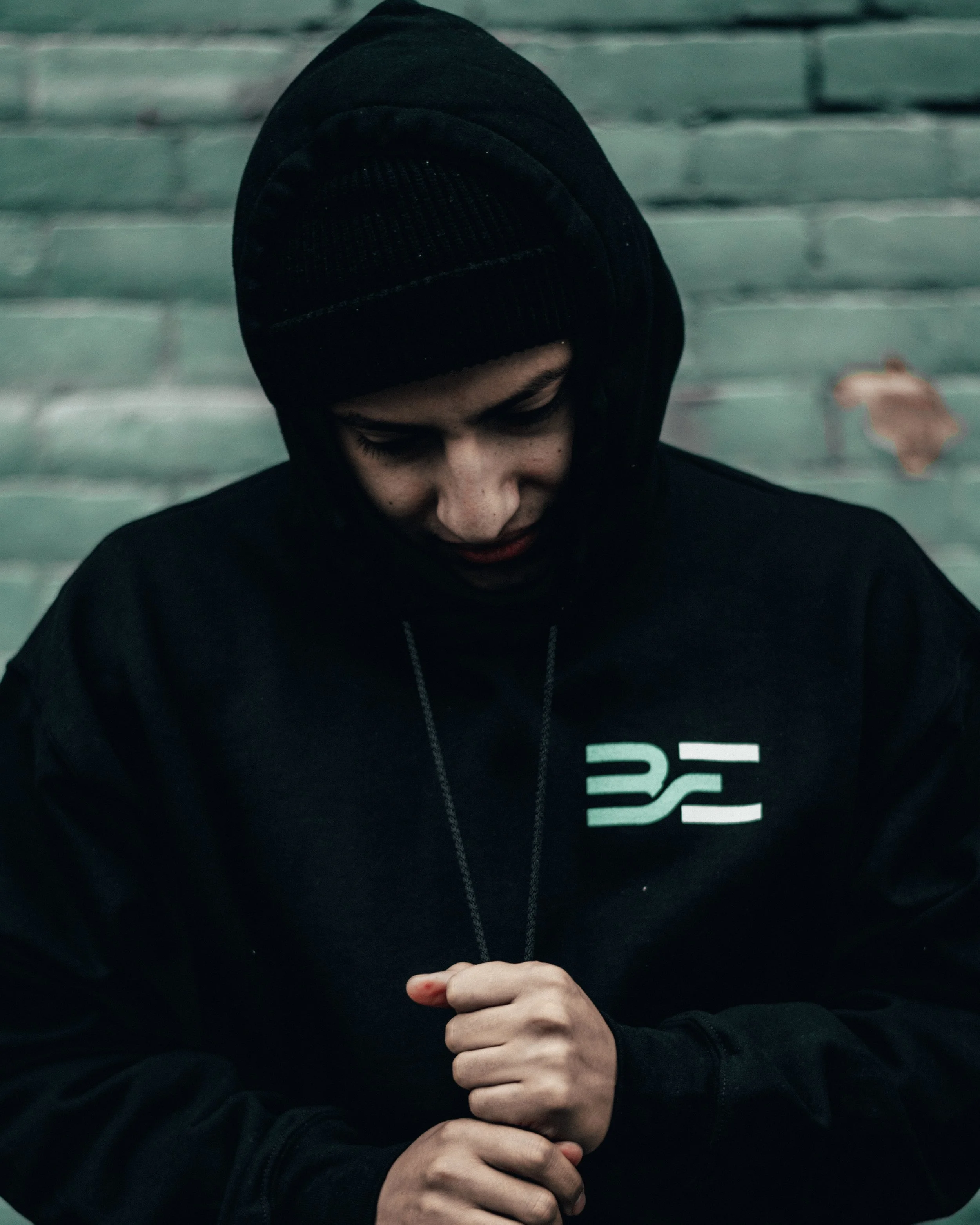 A young man wearing a black hoodie with a logo, hood pulled over his head, looking down, standing against a weathered green brick wall.