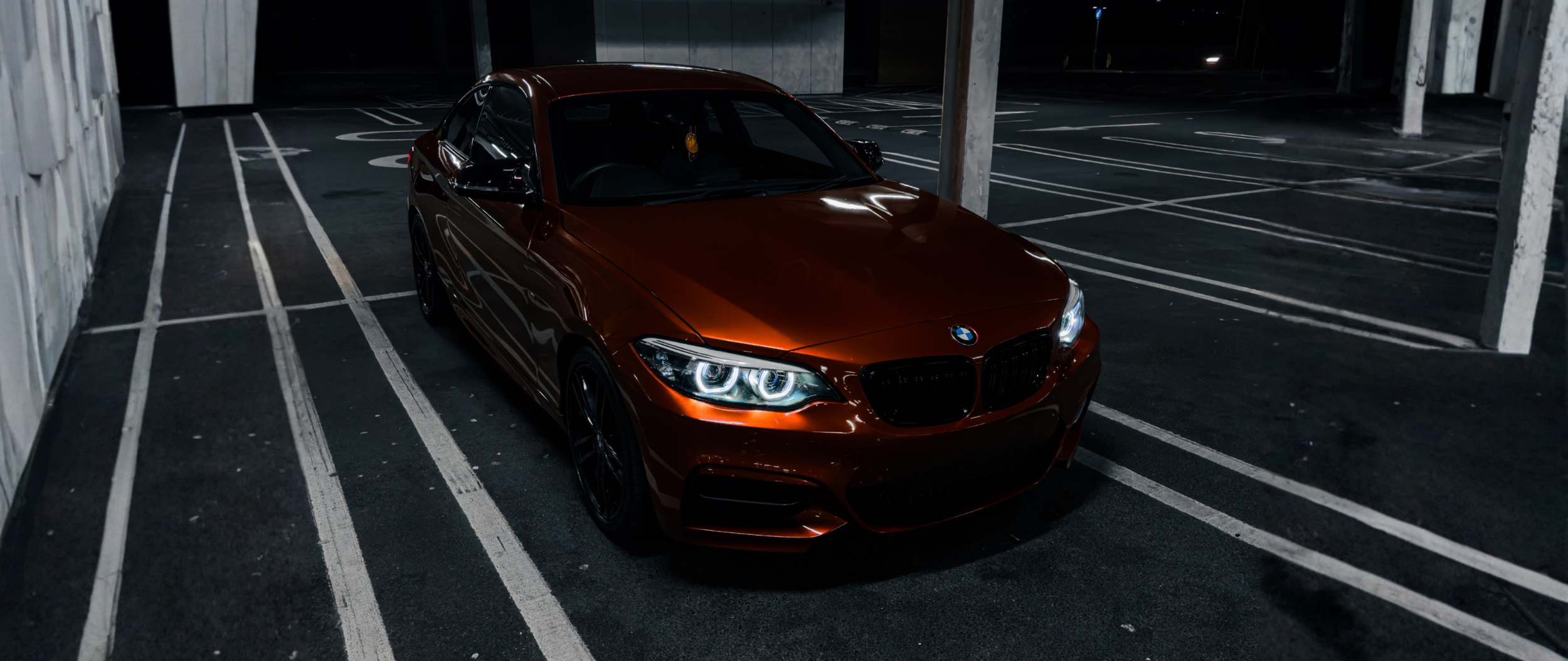 A Sunset Orange coloured BMW sedan parked in an empty, dimly lit parking lot at night.