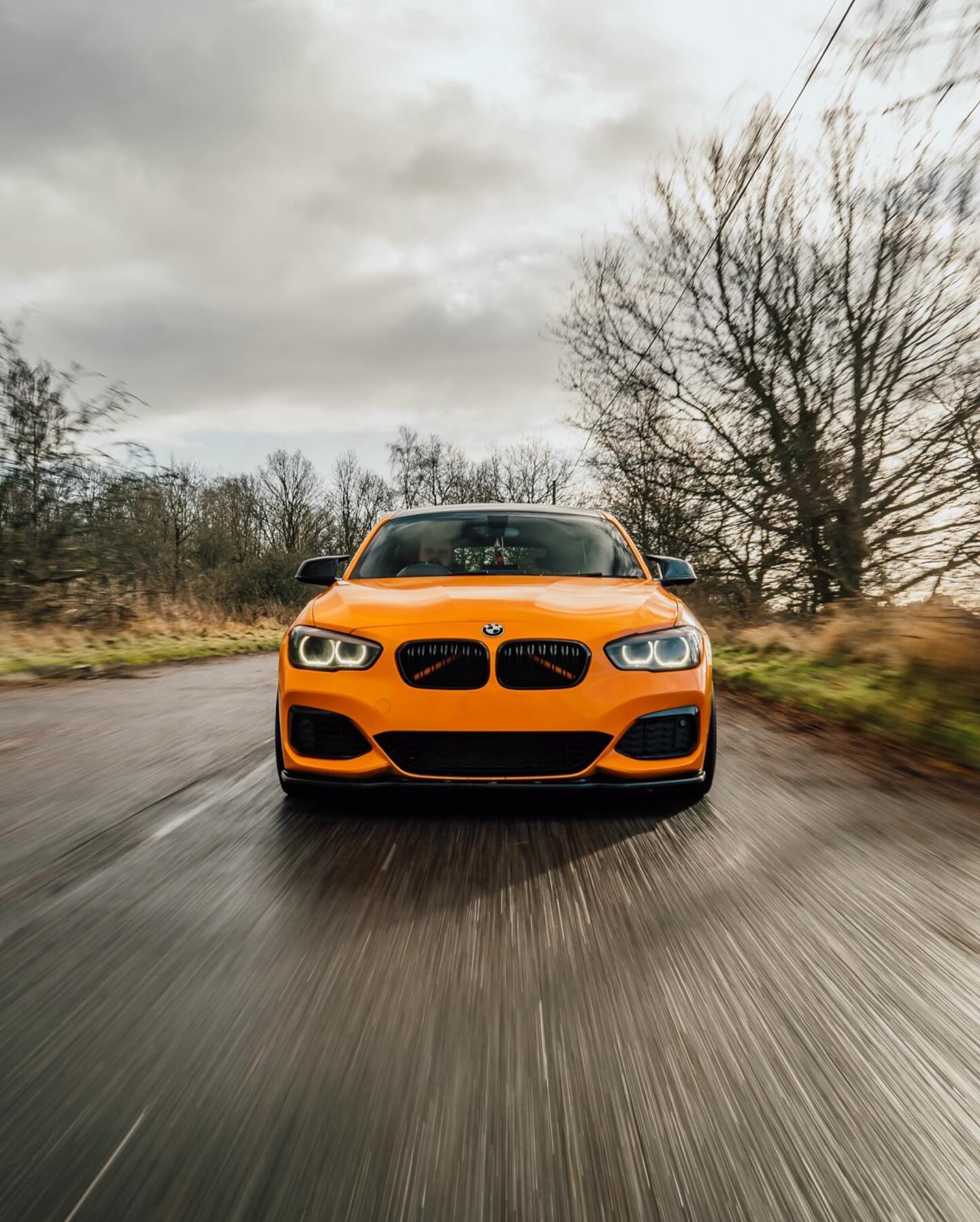 An orange BMW car driving on a countryside road with trees on either side and a cloudy sky overhead.