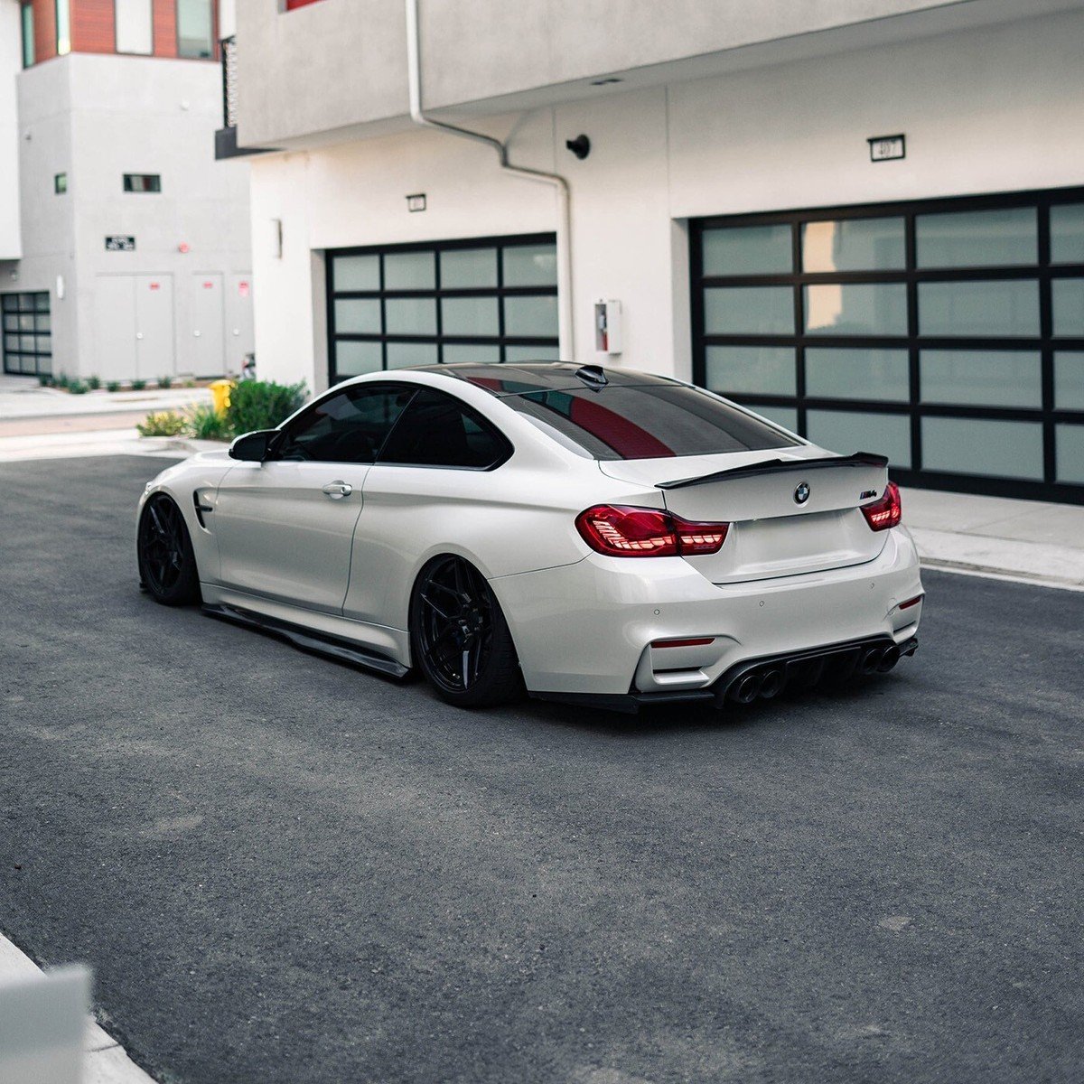 White BMW M4 sports car parked on a driveway in front of modern buildings with garage doors.
