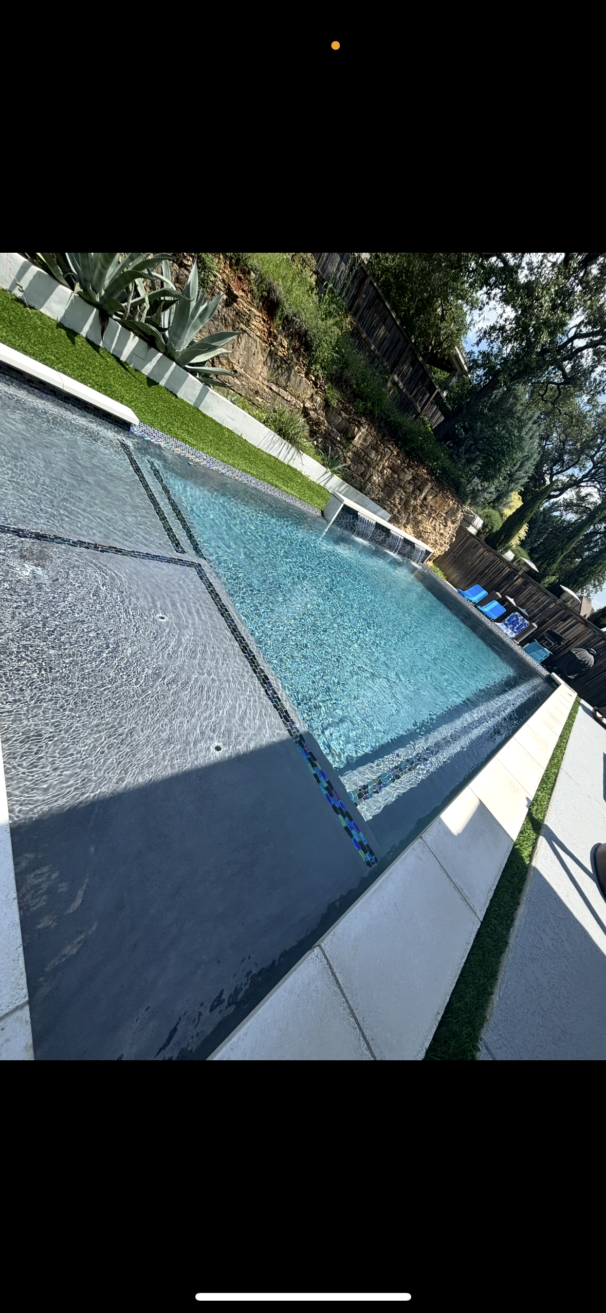 In-ground swimming pool with water features, surrounded by a concrete deck and green lawn, with lounge chairs and trees in the background.