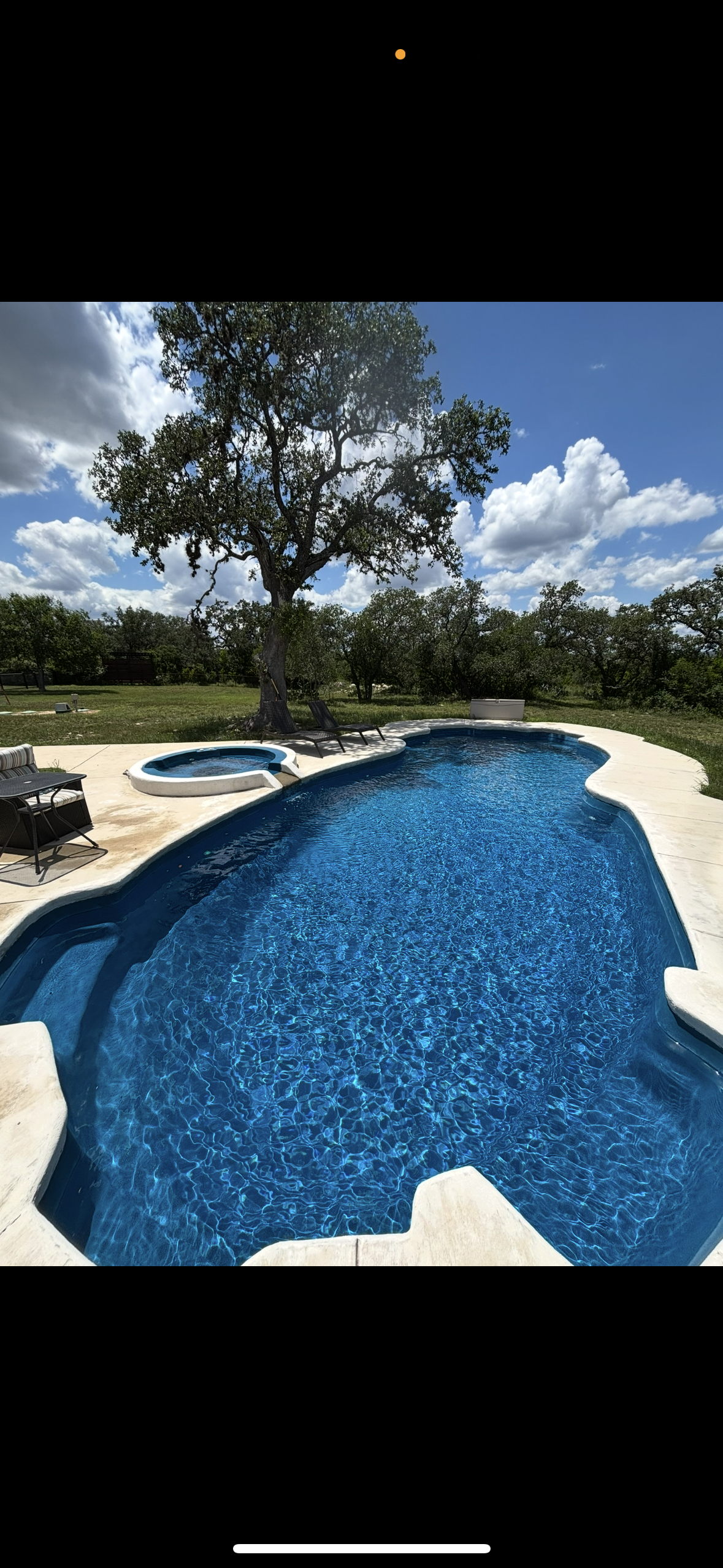 An outdoor swimming pool with a hot tub surrounded by a concrete deck. There are lounge chairs and a tree providing shade near the pool. The sky is partly cloudy with blue sky visible.