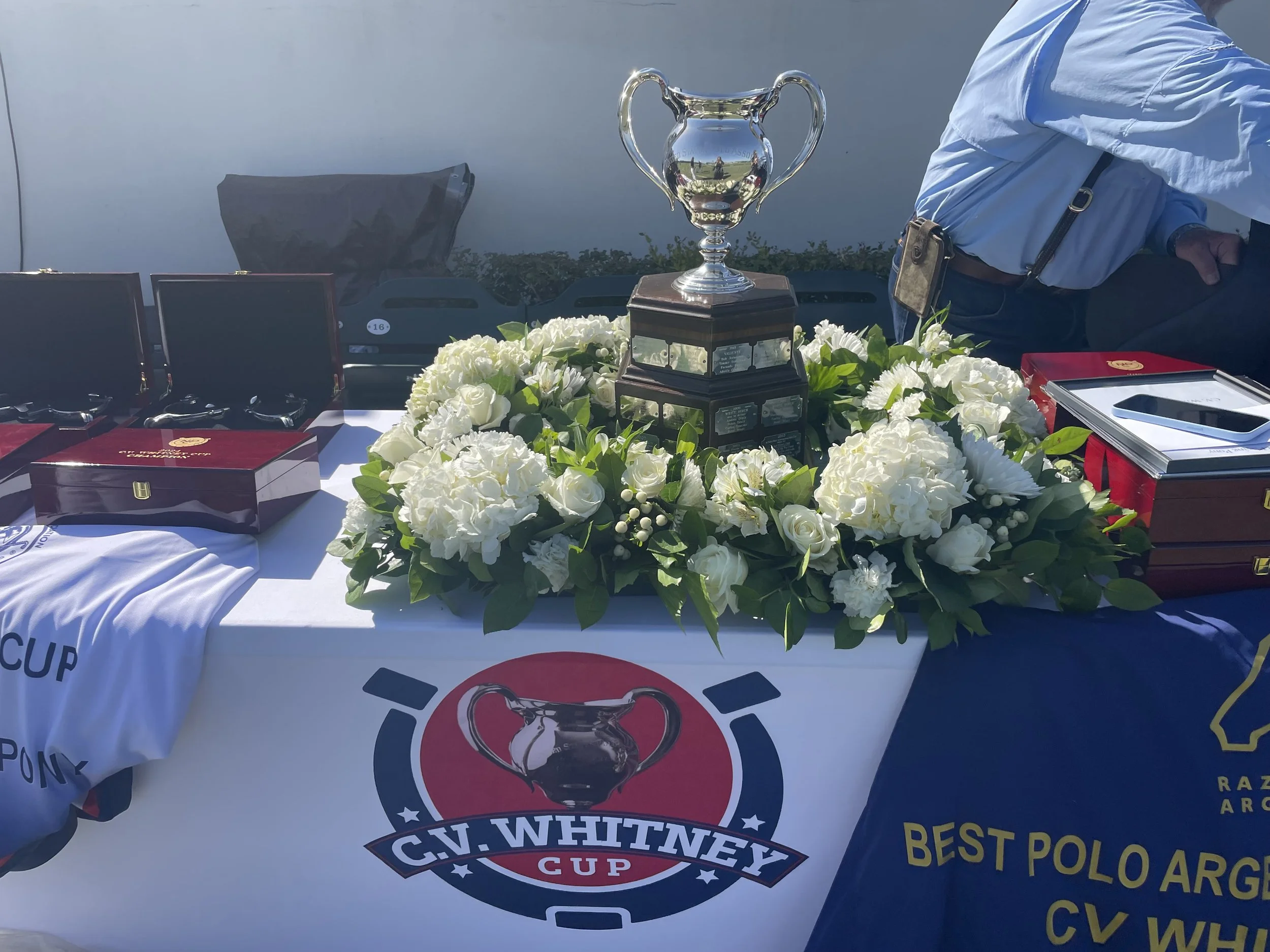 Table with a large trophy, a floral arrangement, boxed medals, and a banner for the C.V. Whitney Cup on a golf course.