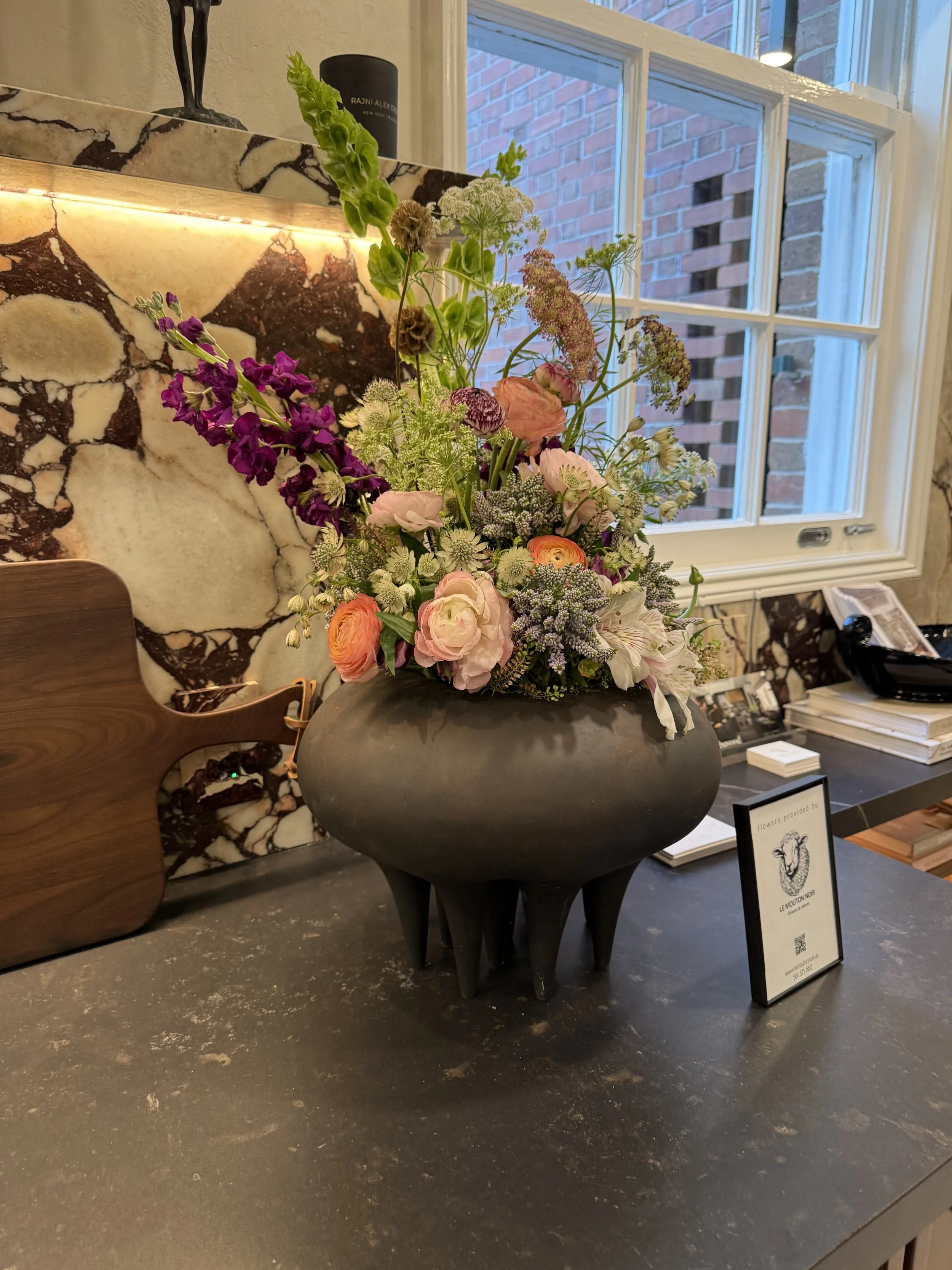 A black, modern vase with four legs filled with a large arrangement of colorful flowers including pink ranunculus, white lilacs, purple snapdragons, and assorted greenery, placed on a marble-topped counter near a window.