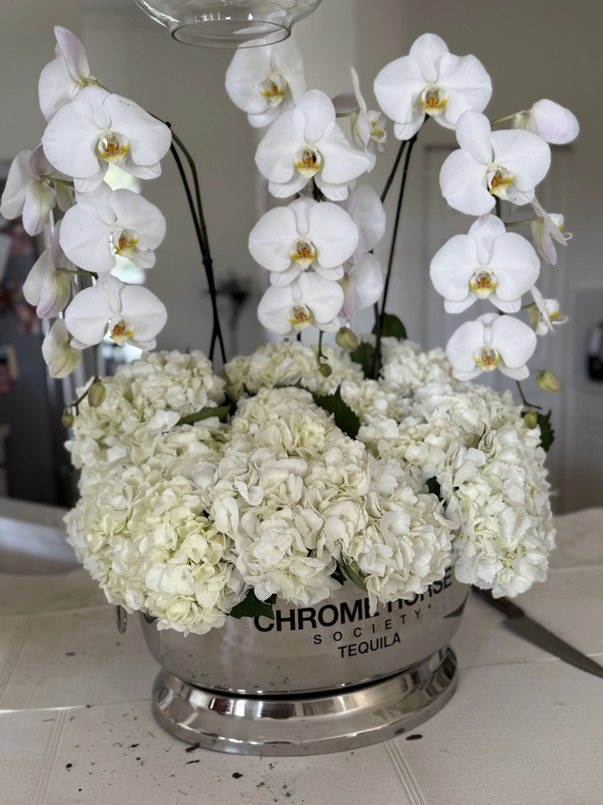 Arrangement of white orchids and hydrangeas in a silver bucket on a table.