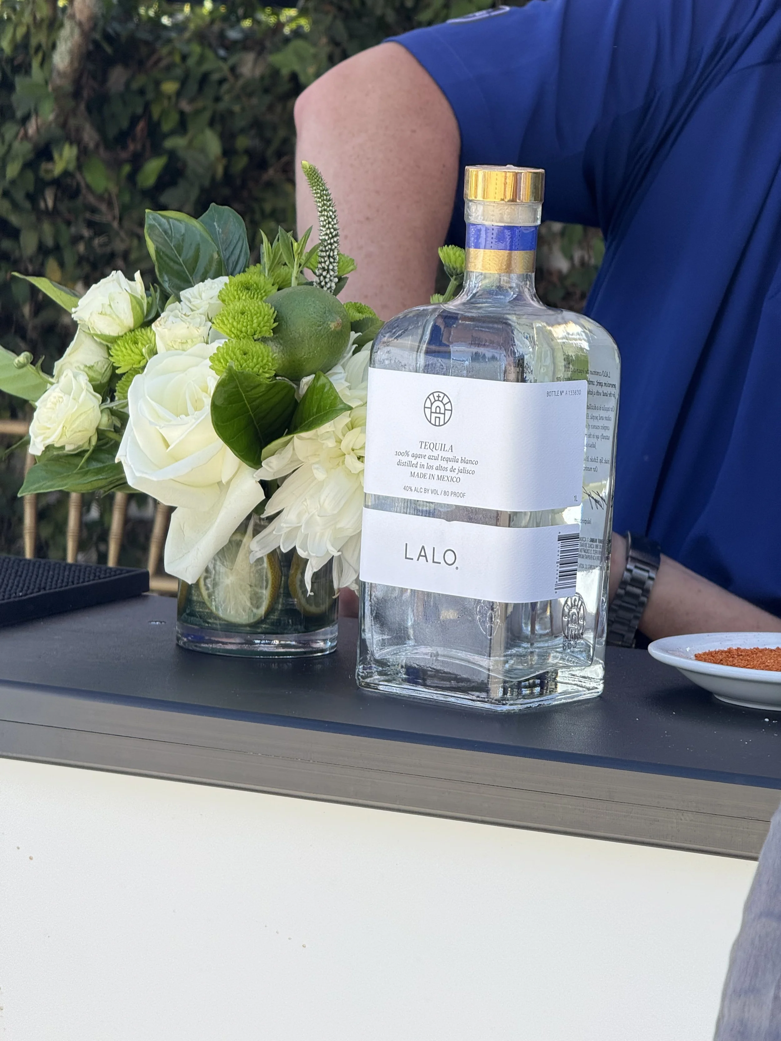 A bouquet of white roses and greenery in a glass vase with lemon slices, a lime, and other accents, next to a large bottle of tequila labeled 'LALO' and a small dish with spices on a black surface.