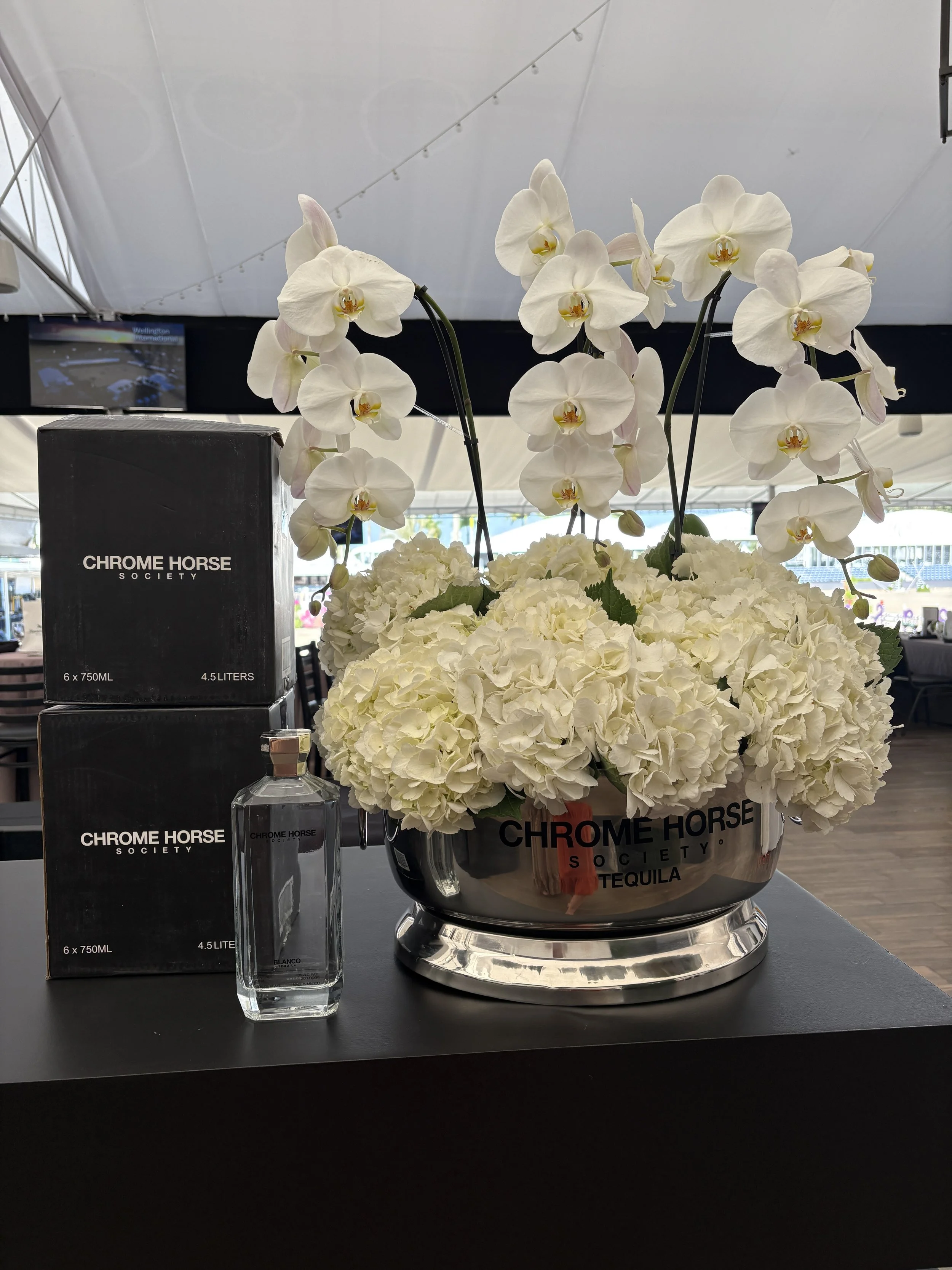 A floral arrangement of white orchids and white hydrangeas in a chrome bowl with 'Chrome Horse Society' written on it, next to boxes of 'Chrome Horse Society' liquor and a bottle of tequila on a black table.