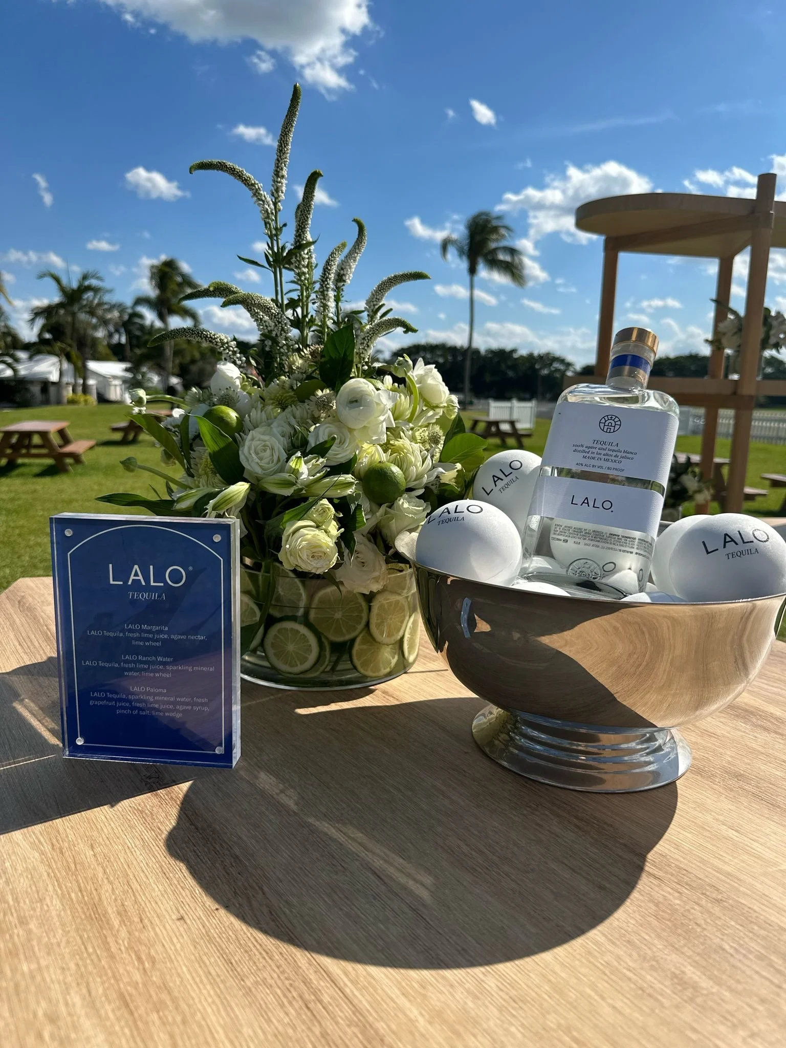 A floral arrangement with white flowers and limes, a bottle of LALO tequila, branded balls, and a blue menu card on a wooden table outdoors.