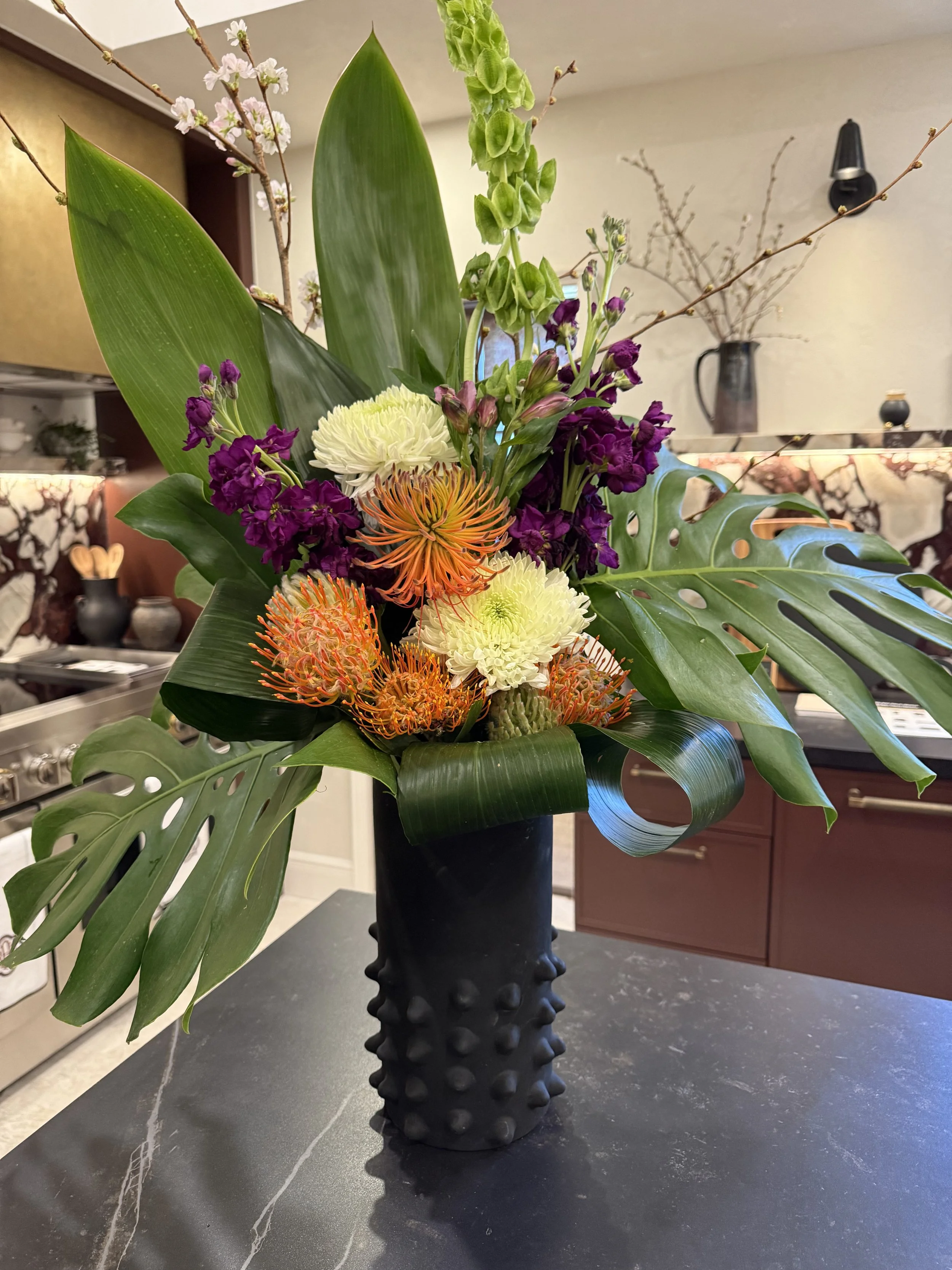 A vibrant floral arrangement featuring large monstera leaves, purple delphinium, white chrysanthemums, orange pincushion protea, green snapdragons, pink cherry blossoms, and curly lily grass in a textured black vase on a dark kitchen countertop.