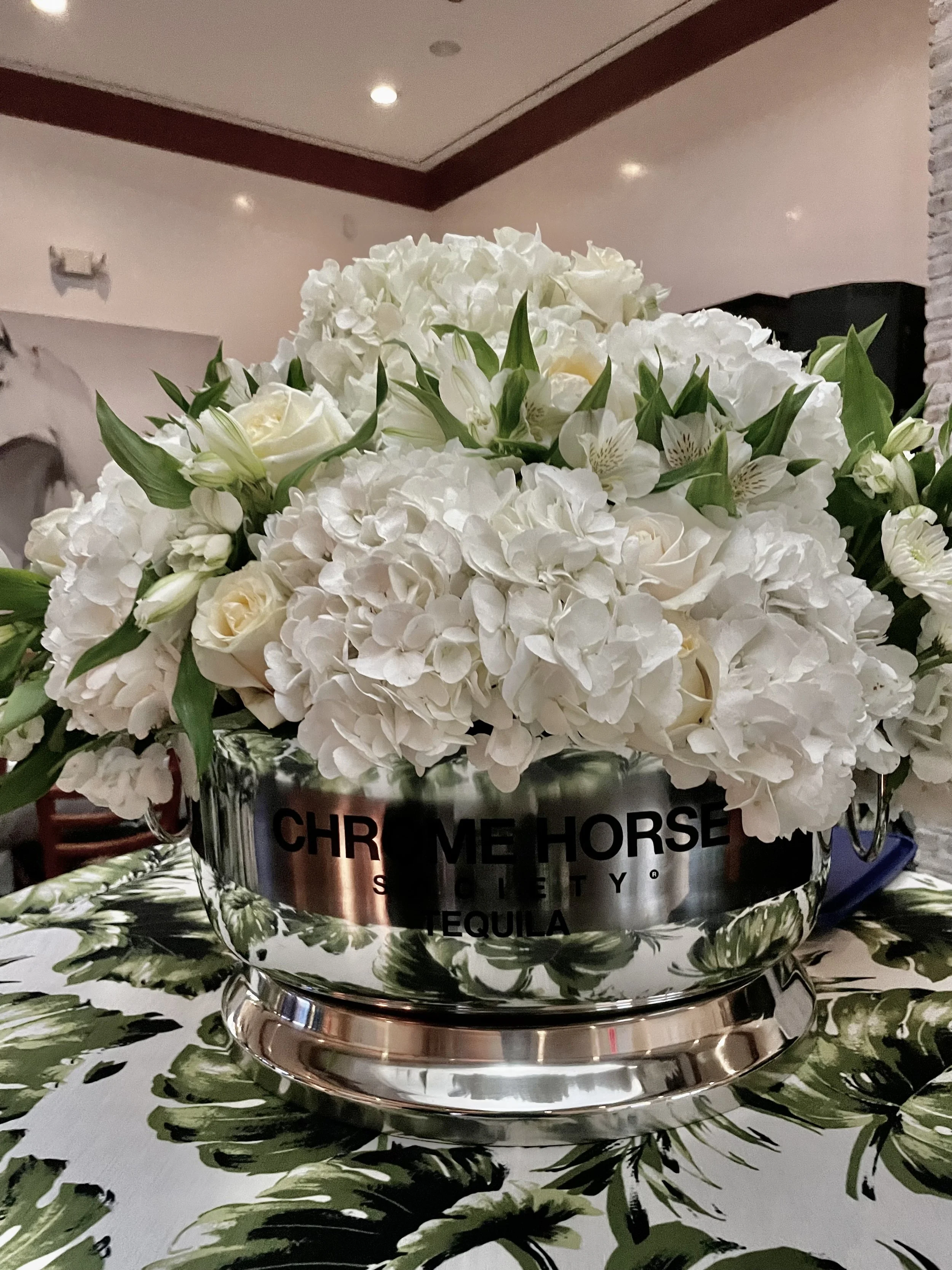 A silver flower arrangement container labeled 'Chrome Horse' with white hydrangeas, roses, and greenery on a table with a patterned tablecloth.