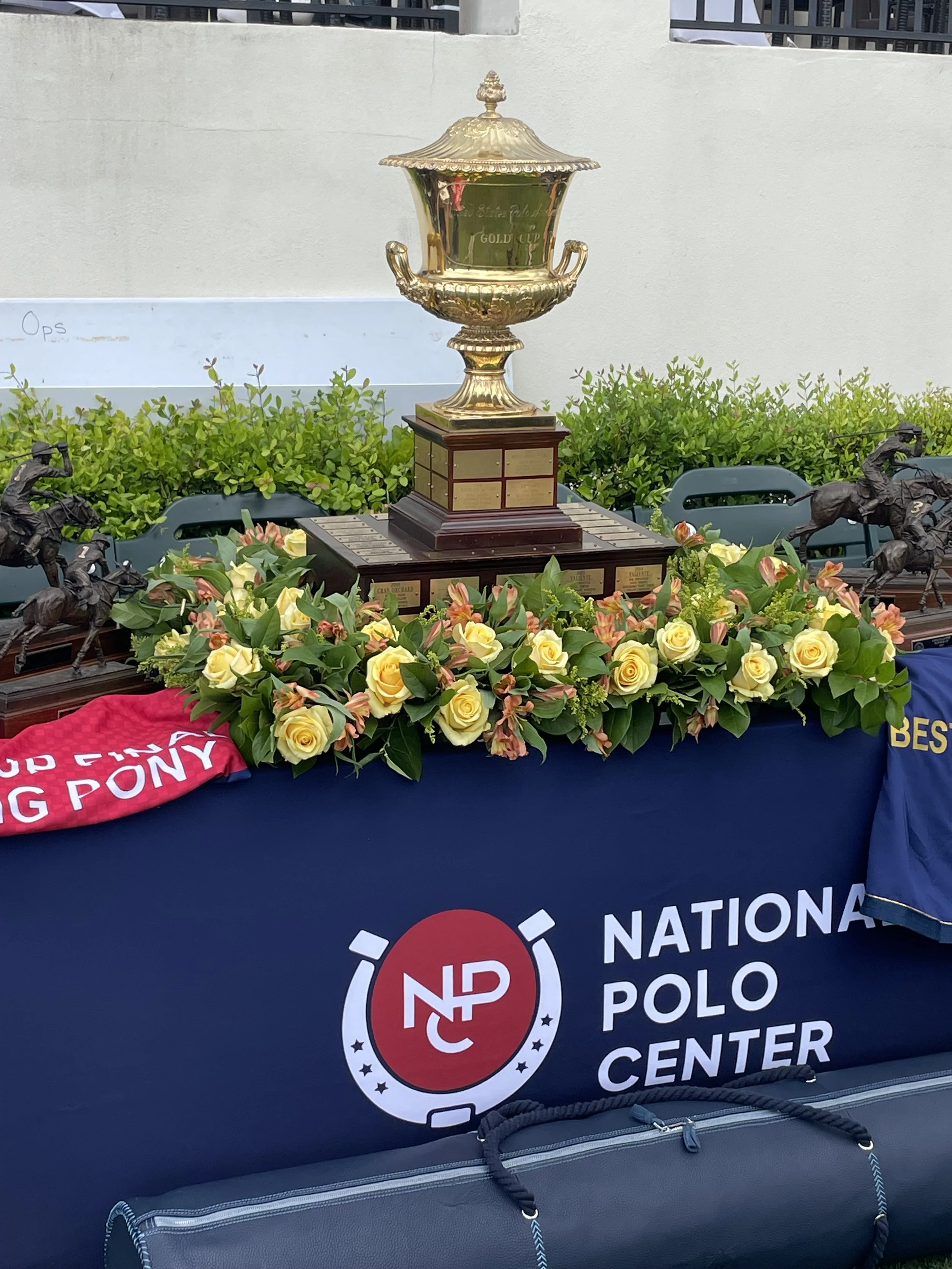 A large gold trophy with handles on a wooden base, surrounded by a floral arrangement of yellow roses and pink flowers, displayed on a table covered with a dark blue cloth with the words 'National Polo Center' and the logo. There are bronze horse fig