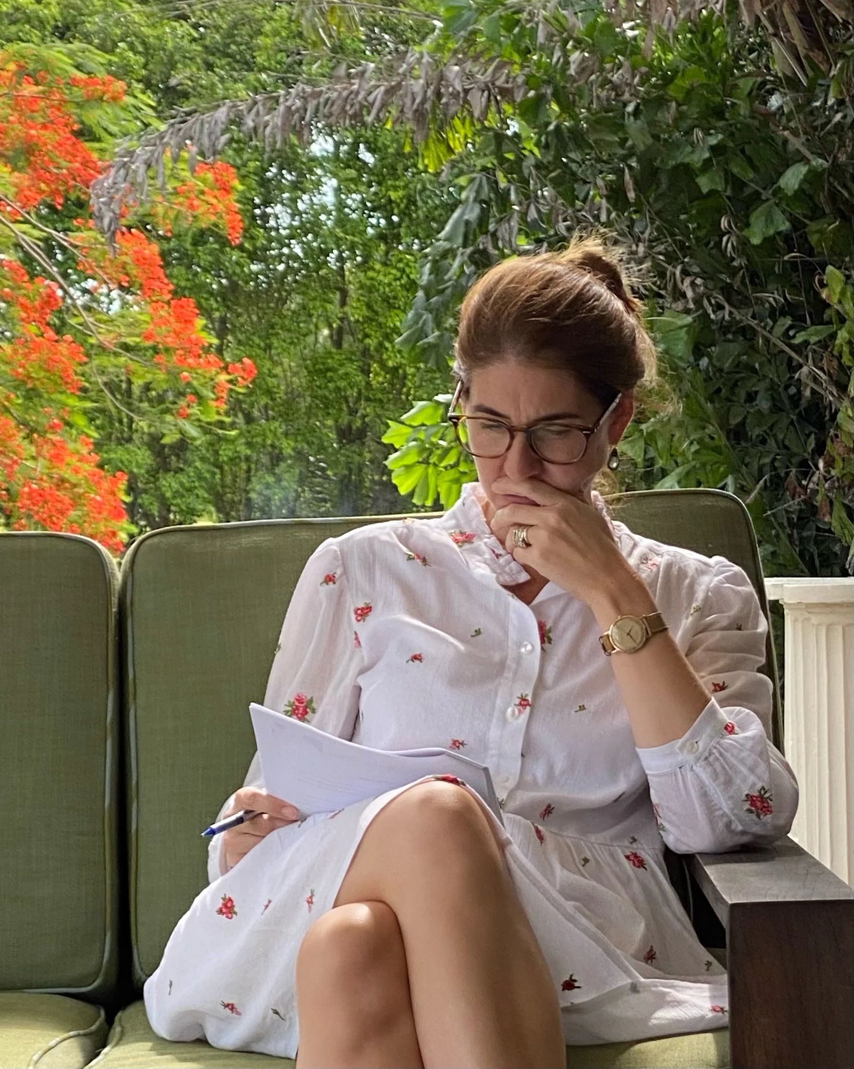 A woman sitting outside on a green couch, wearing glasses and a white dress with a floral pattern, appears to be deep in thought while holding a notepad and pen, with lush greenery and red flowers in the background.