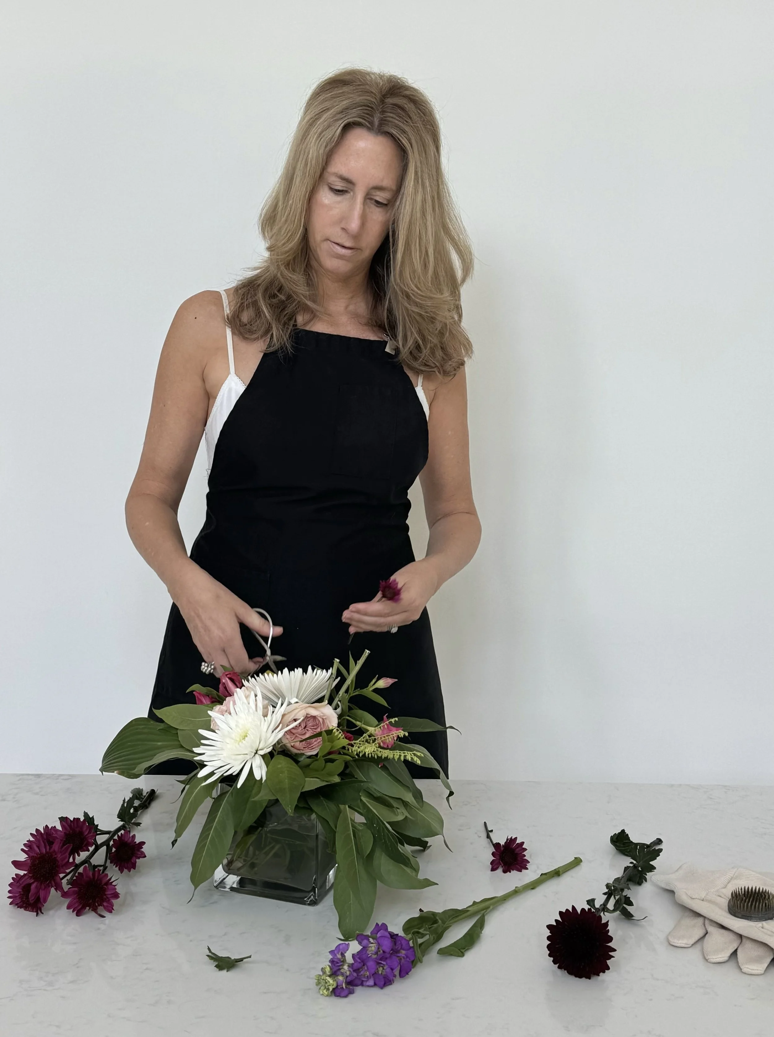 Woman arranging a colorful flower bouquet on a white table, with flowers and gardening tools around her, against a plain white background.