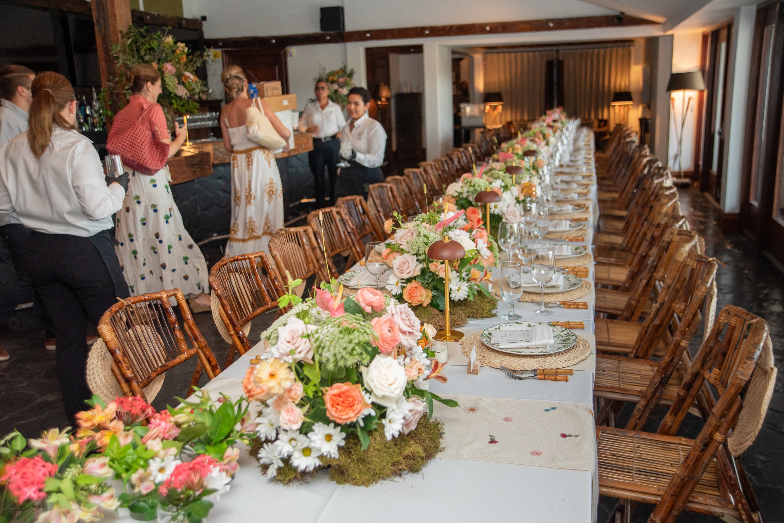 A long dining table set for a celebration with floral centerpieces, place settings, and decorative lamps, in a warmly lit indoor venue with people preparing or attending the event.